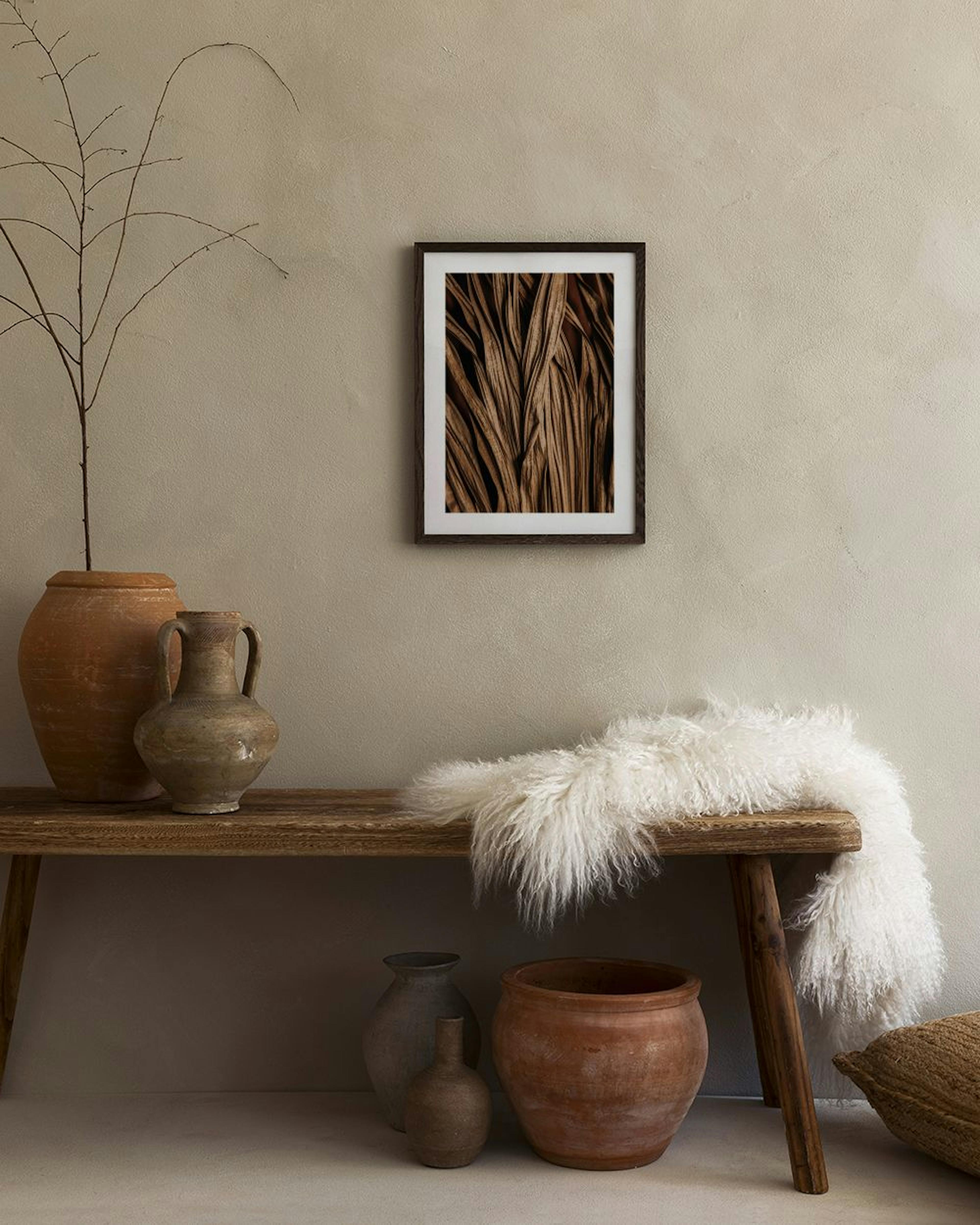 A print of dried palm fronds in brown tones hanging above a cabinet with three vases and two books.