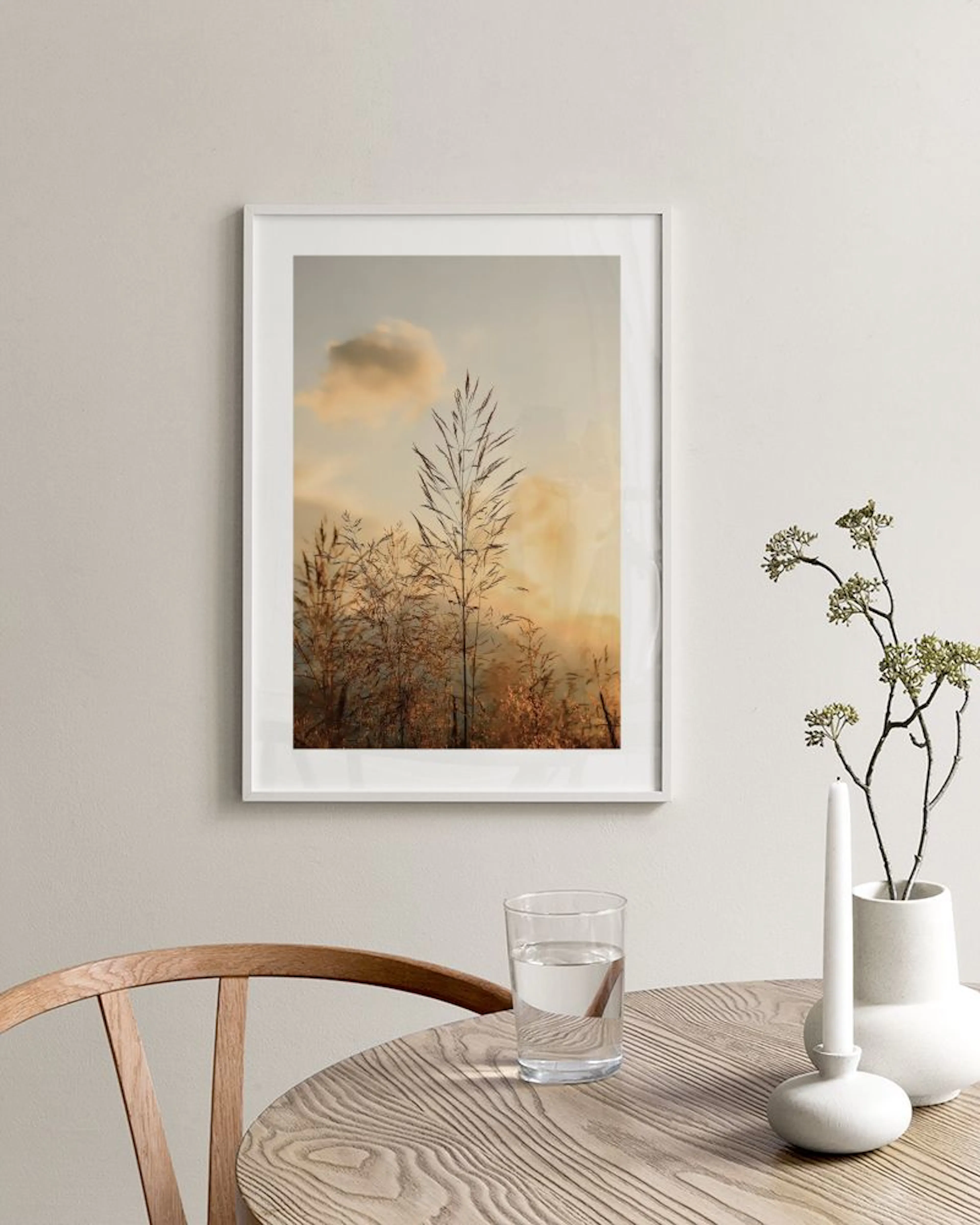 A nature print of tall golden grass with a soft orange and blue sky, displayed in a minimal room above an open book and decorati