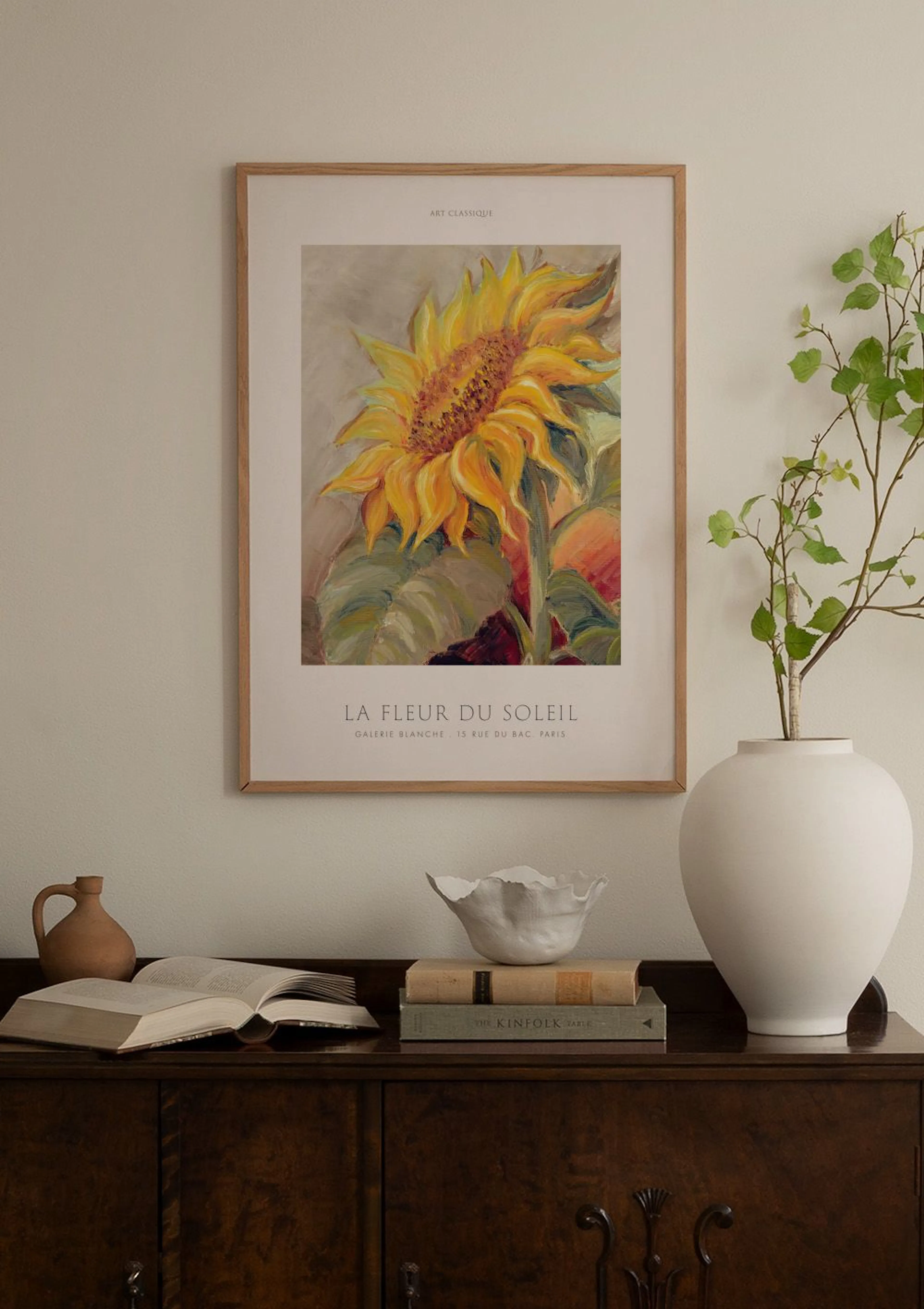 A sunflower print with bold yellow petals and green leaves, hanging above a white cabinet with minimalist vases and books.