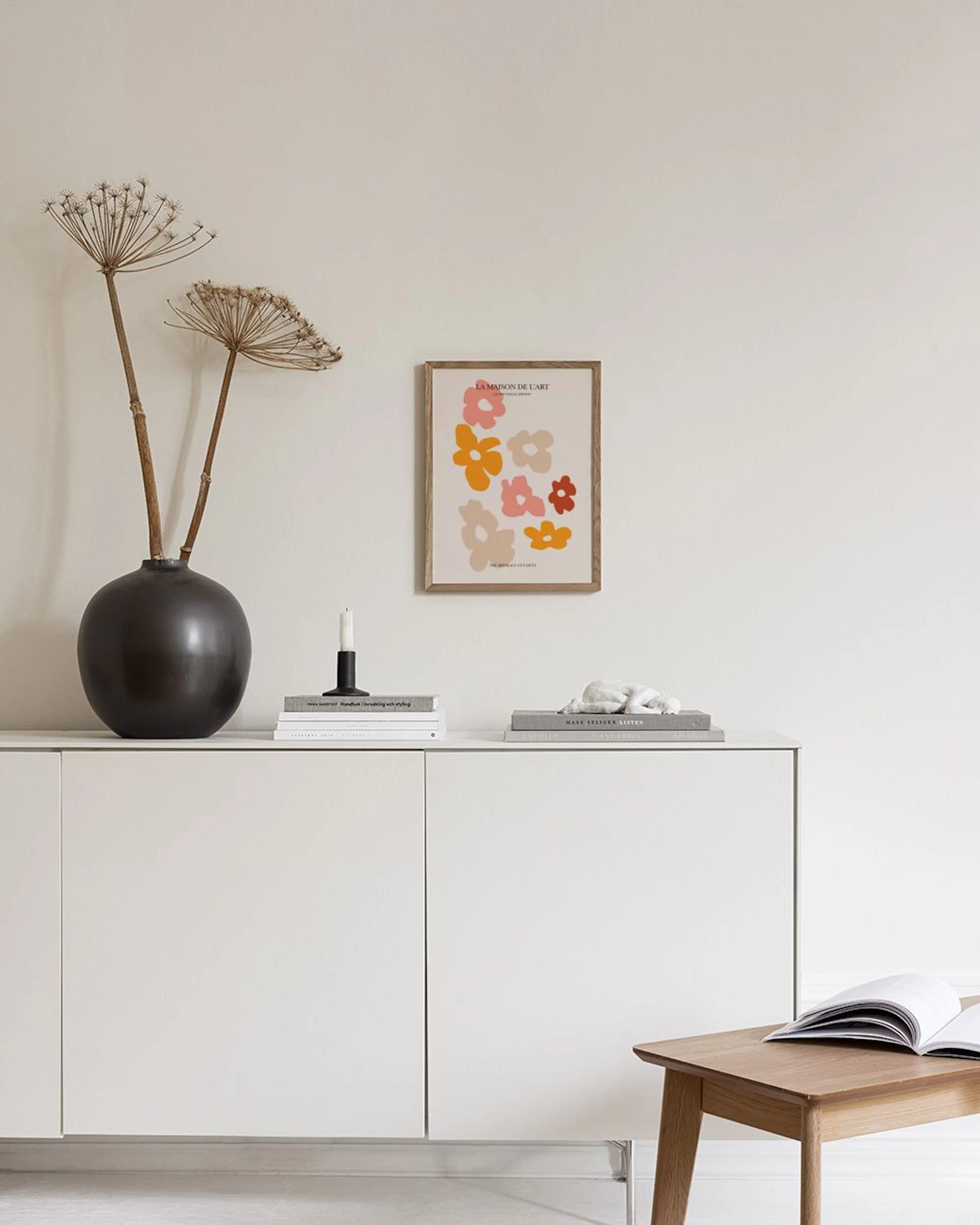 Póster de flores abstractas en tonos naranja, rosa y beige, enmarcado sobre un mueble blanco con jarrones y libros.