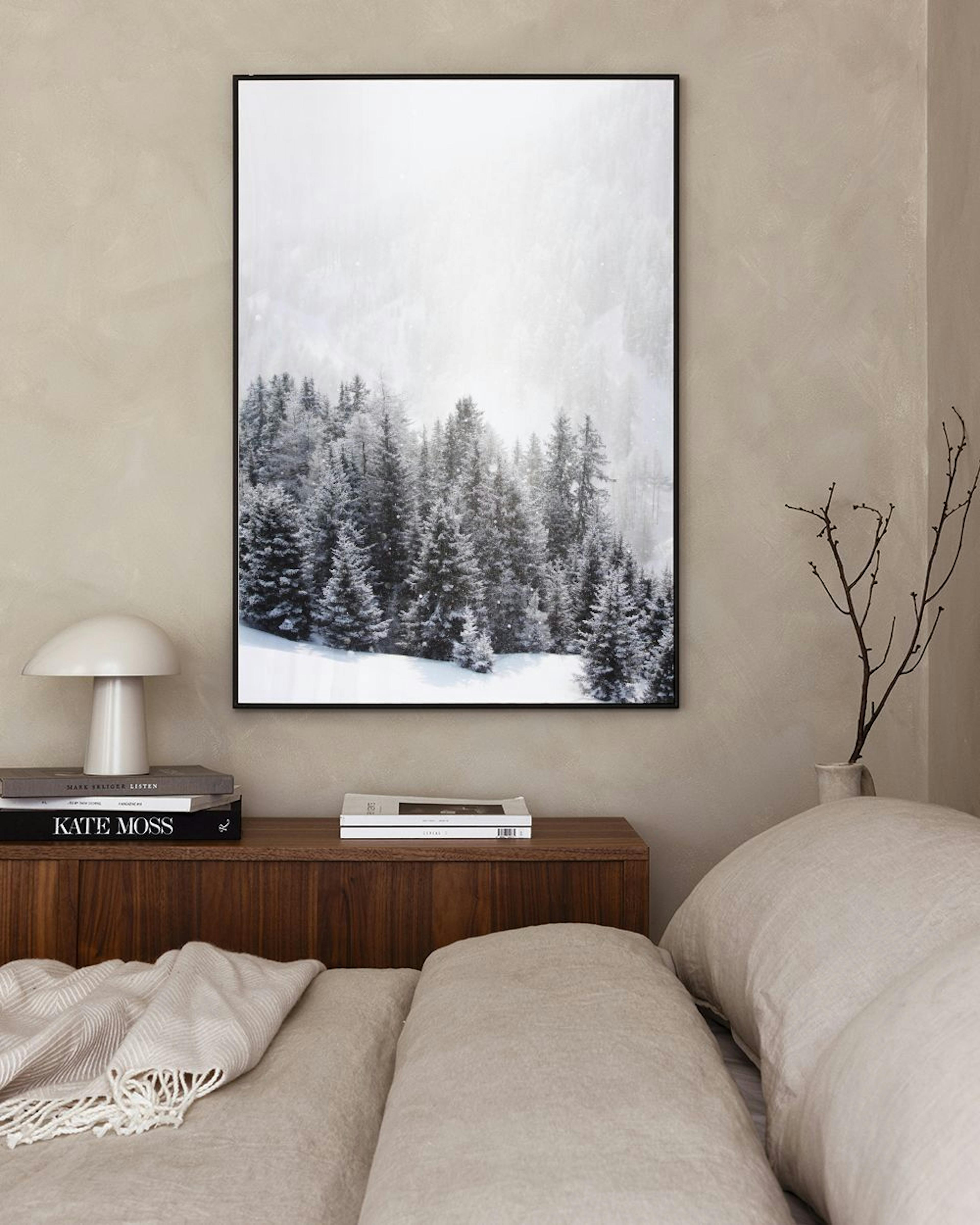 A print of a snow-covered forest of pine trees, with light snow falling, hanging above a bed with a book.