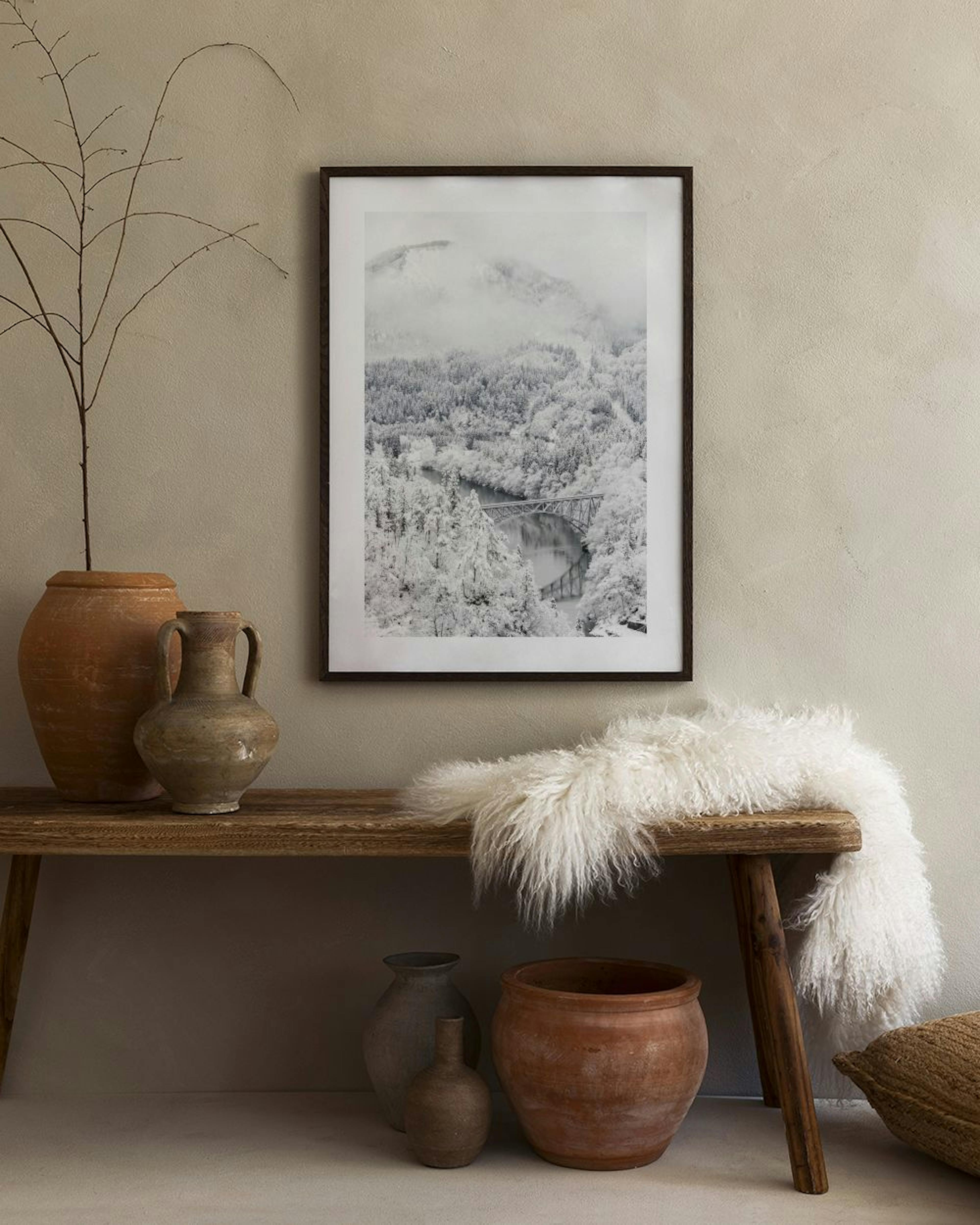 Póster de paisaje nevado con montañas, bosque y un puente sobre un río, colgado en una pared clara sobre una mesa auxiliar.