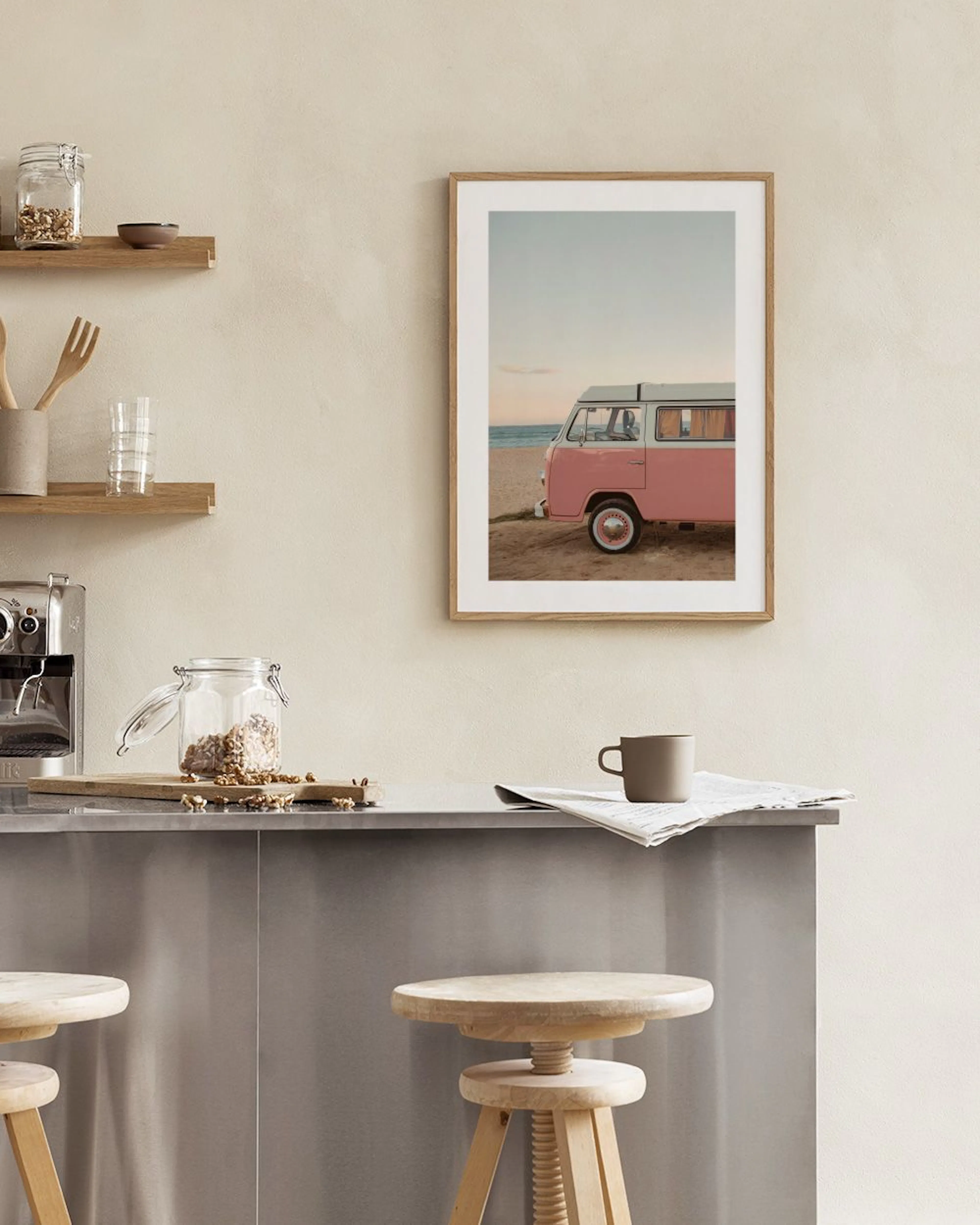 A print of a pink vintage camper van on a sandy beach, hanging above a white console with a glass vase and open book.