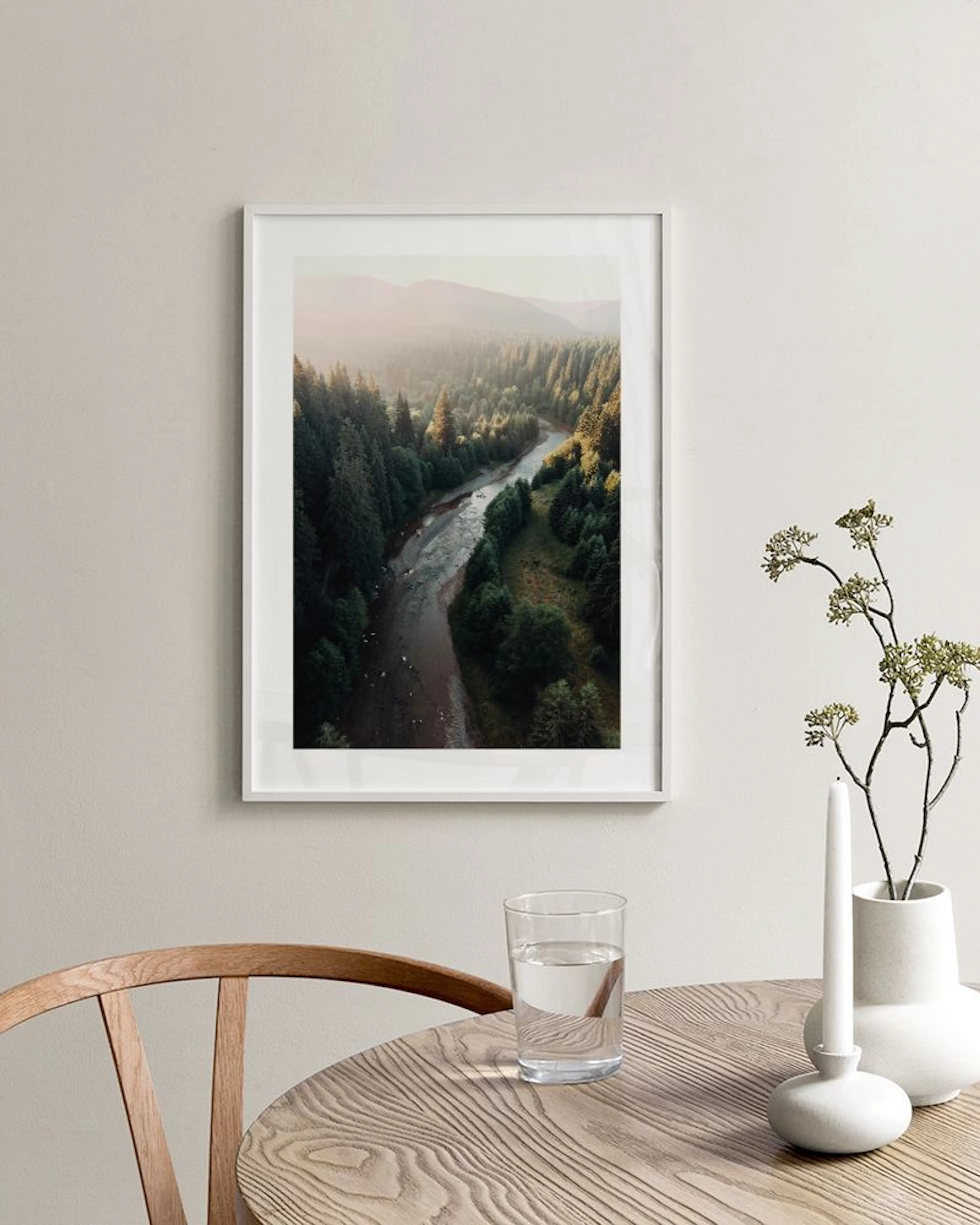 Póster de un río serpenteante entre un denso bosque de pinos y montañas, colgado sobre un mueble blanco con un jarrón y libros.