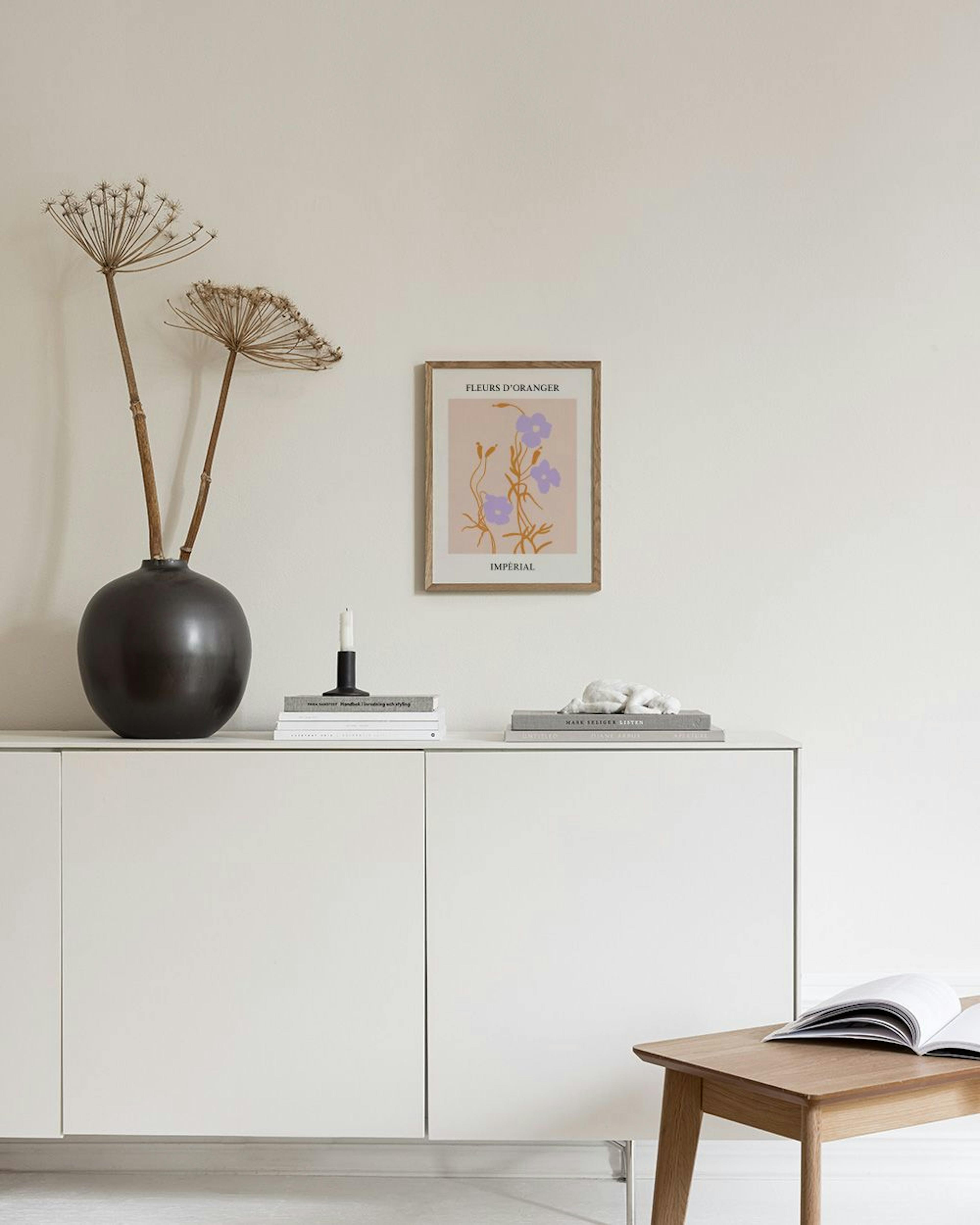 Poster con fiori viola stilizzati e steli arancioni su sfondo beige, appeso sopra un tavolino con vasi bianchi e libri.
