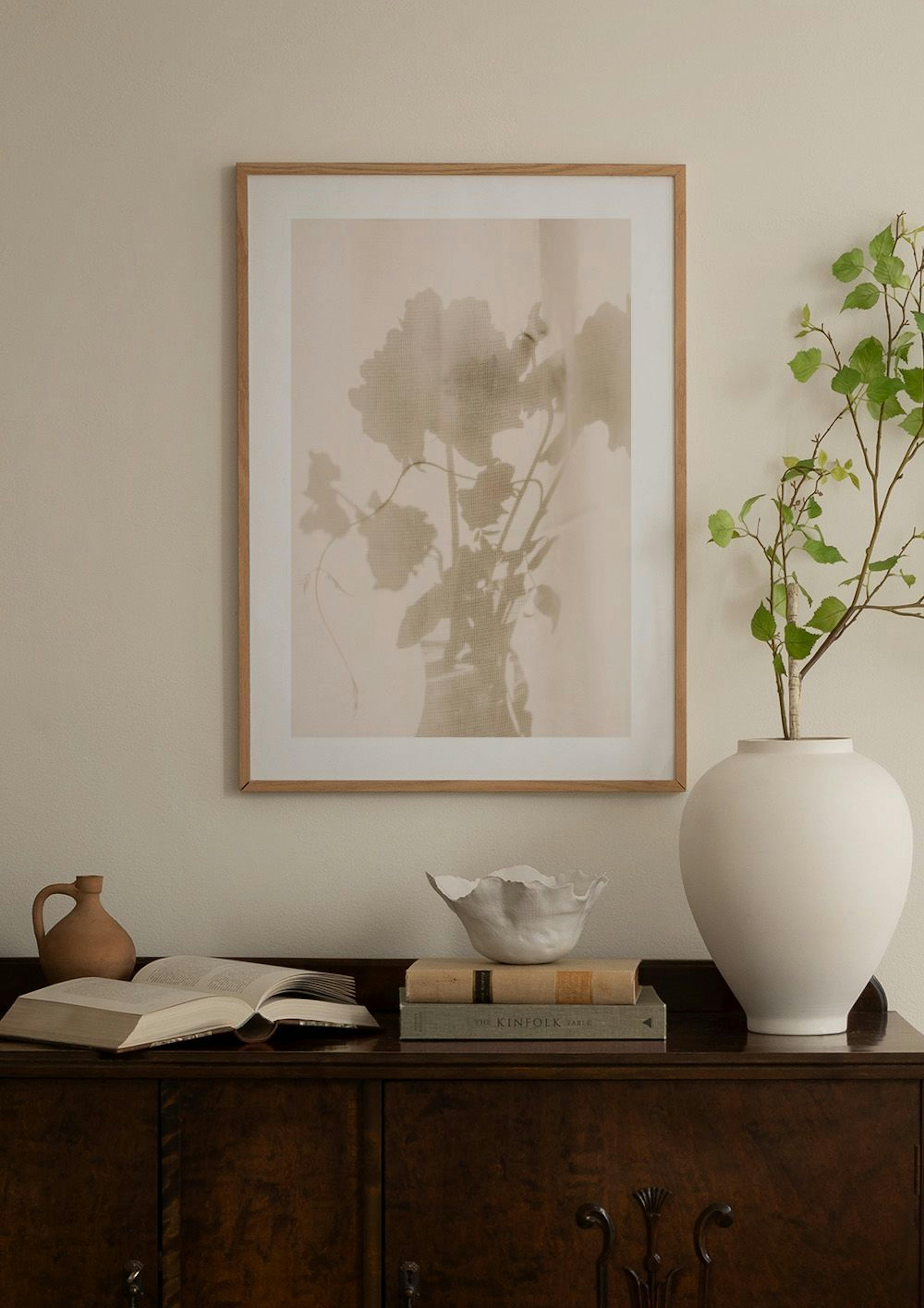 Póster beige con siluetas florales, colgado sobre un mueble blanco con un jarrón de cristal y libros abiertos.