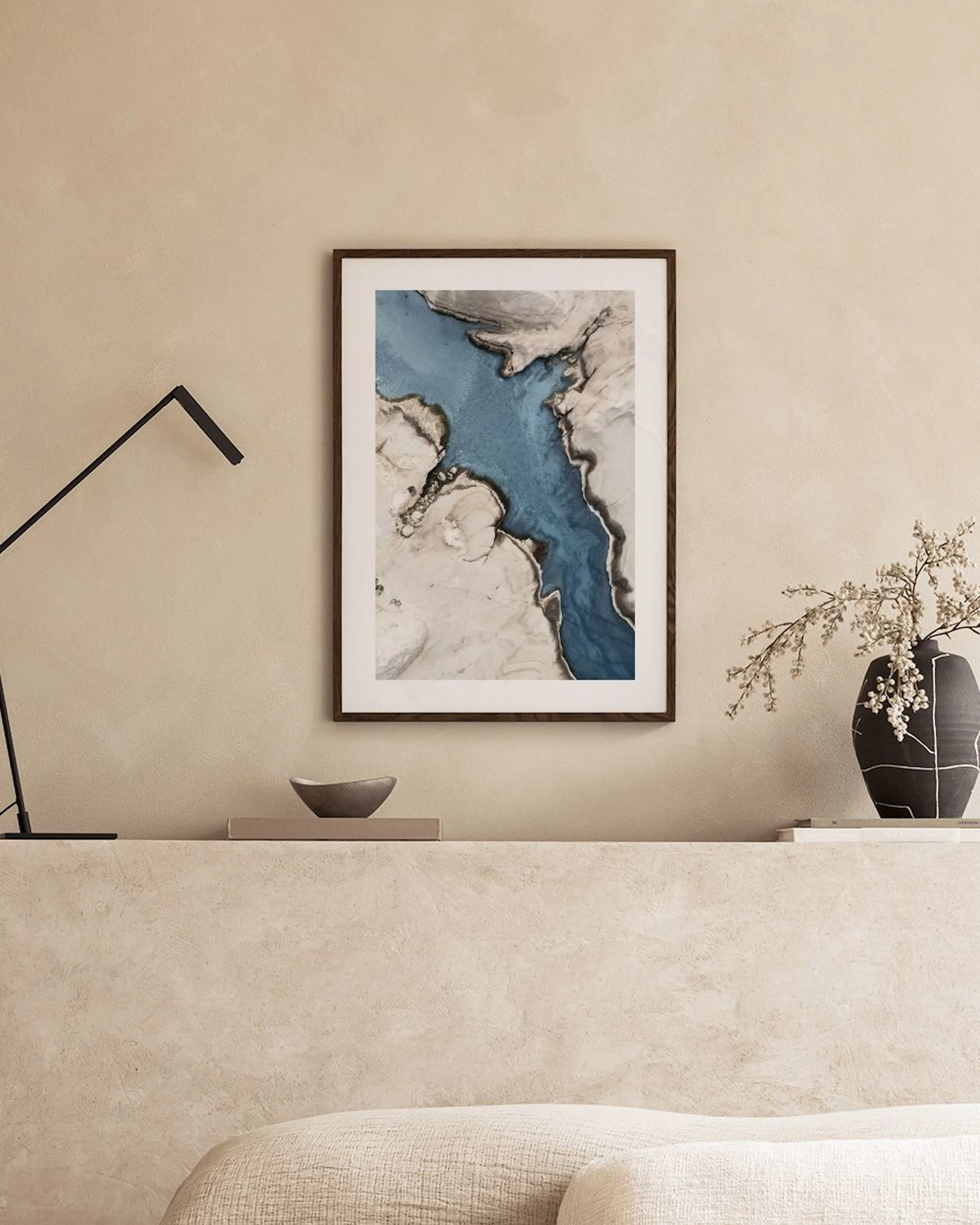 A print of a winding blue river through abstract white and beige land, hanging above a rustic wooden bench with vases.