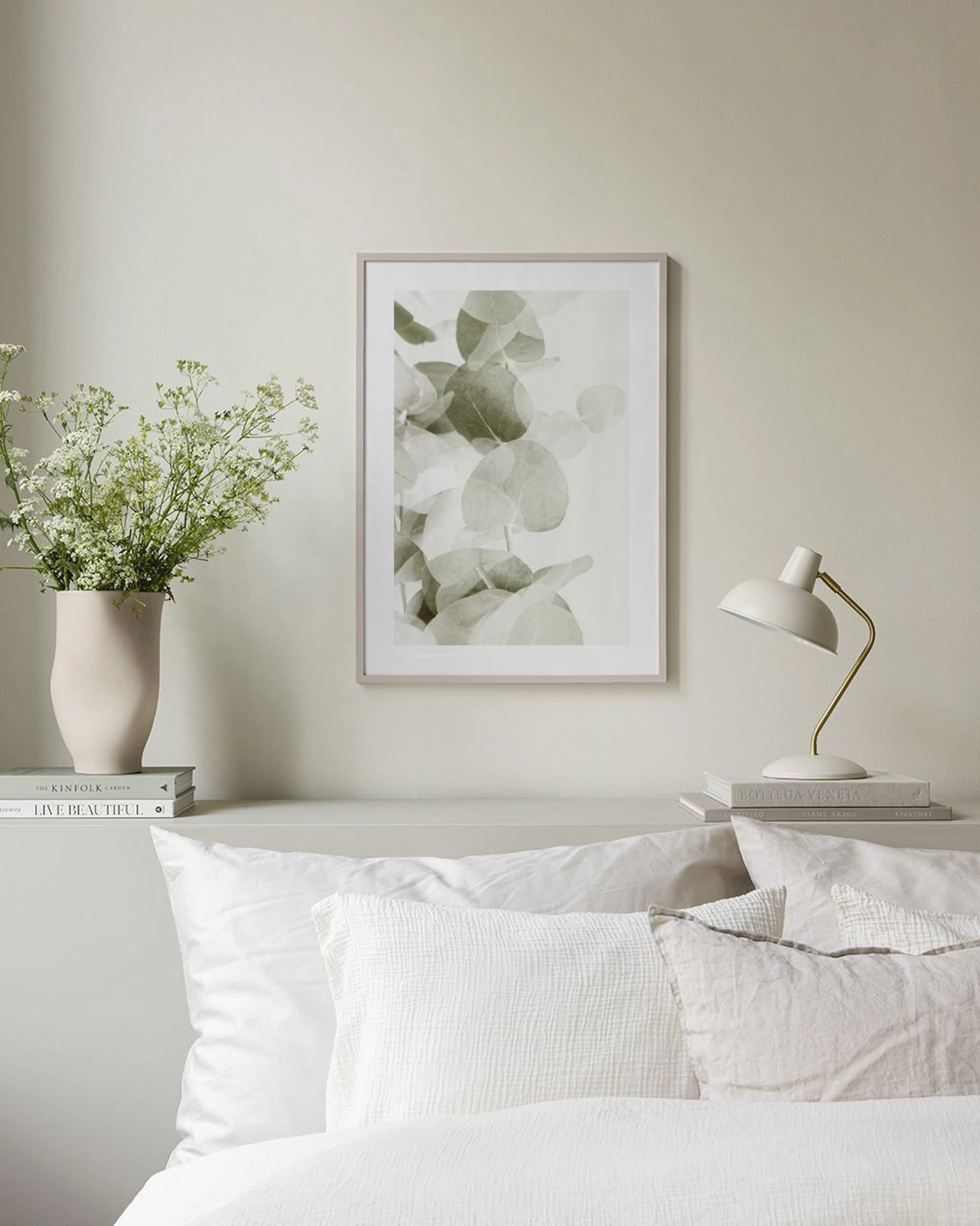 Póster de hojas de eucalipto en tonos verdes y blanco, colgado sobre un mueble blanco con un jarrón y un libro abierto.