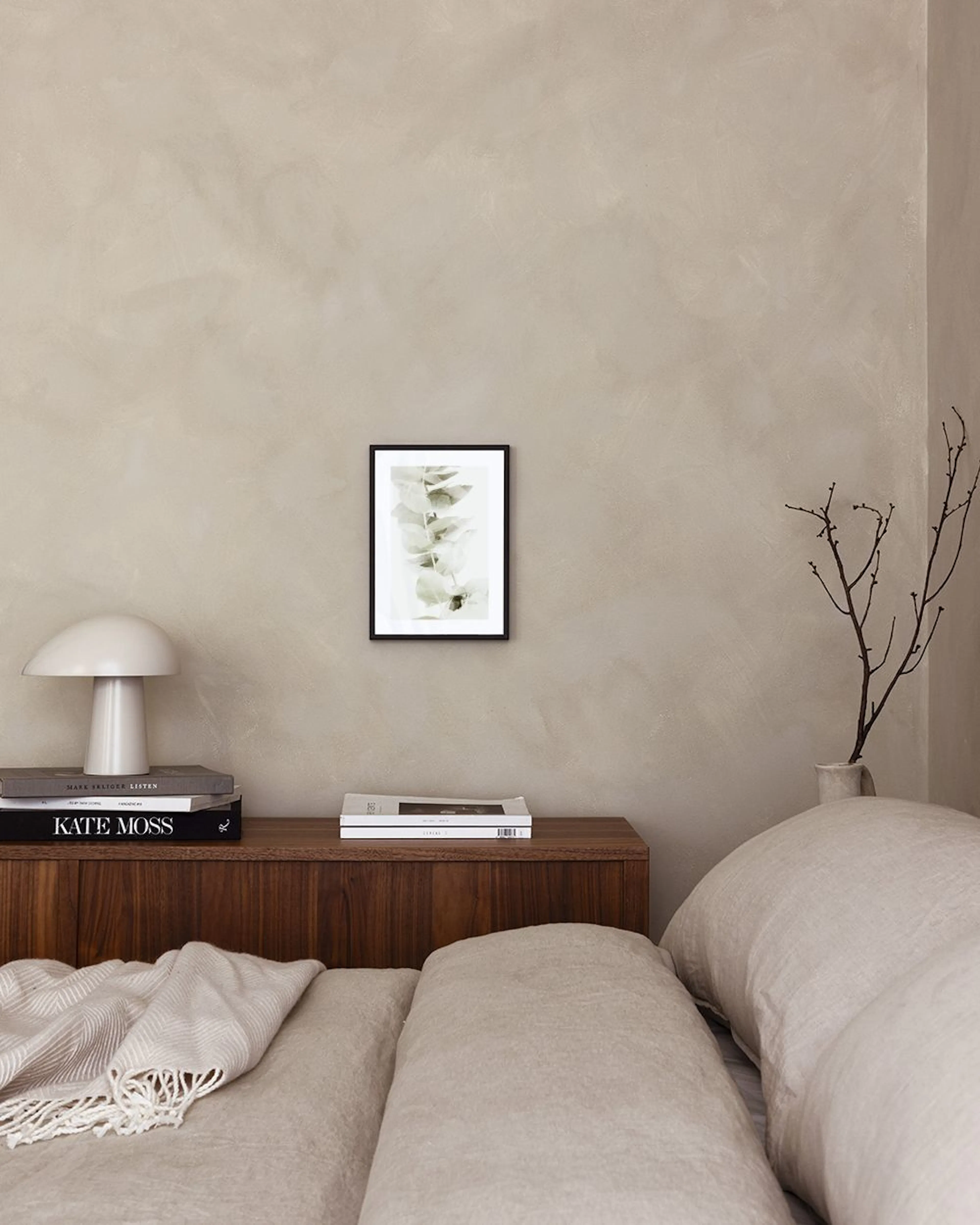 Een poster met eucalyptusbladeren in zachte groentinten, hangend boven een wit marmeren bijzettafel met twee vazen en boeken.