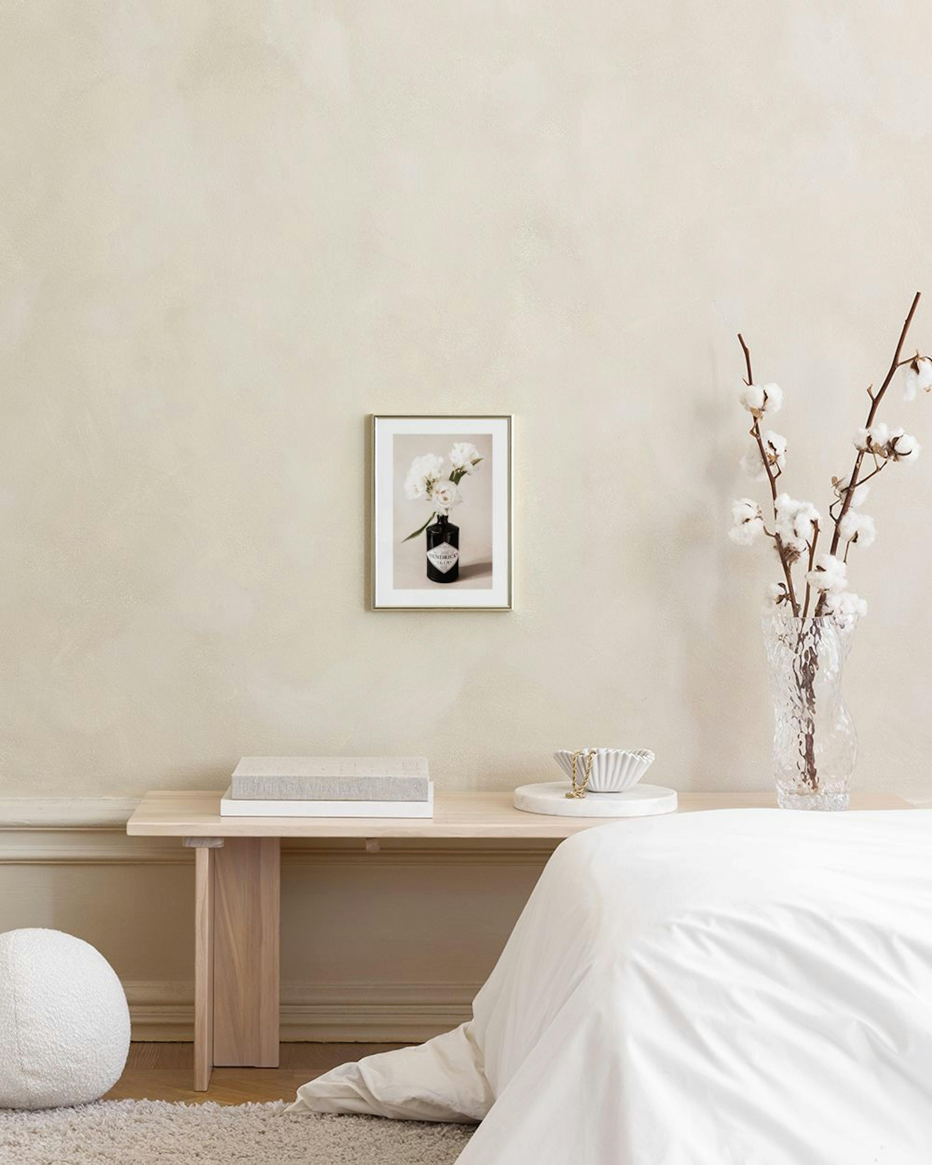 A print of white flowers in a black Hendricks gin bottle, hanging above a brown cabinet with decorative vases and books.