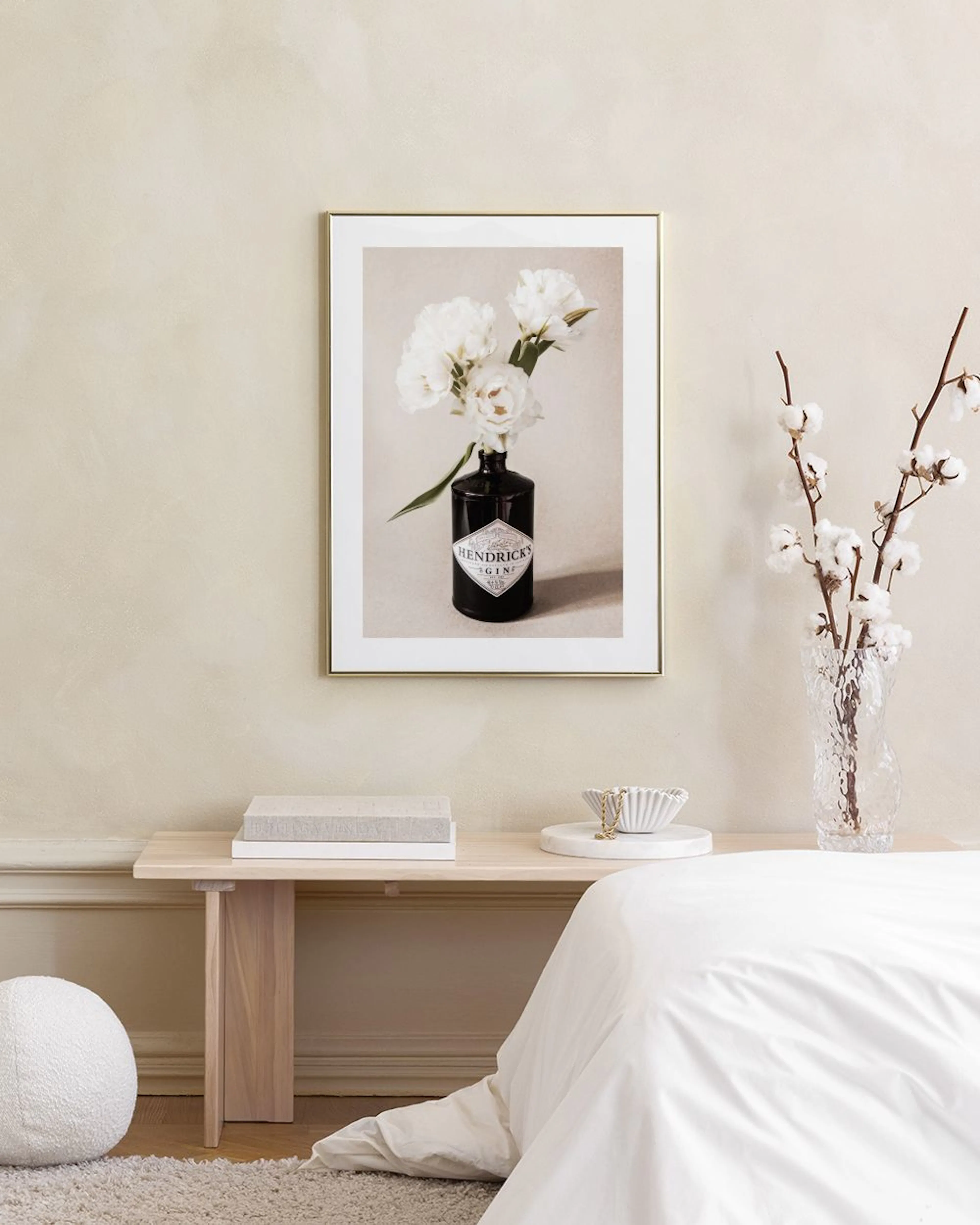 Plakat med hvide blomster i en sort Hendricks ginflaske, hængende over et brunt skab med vaser og bøger.
