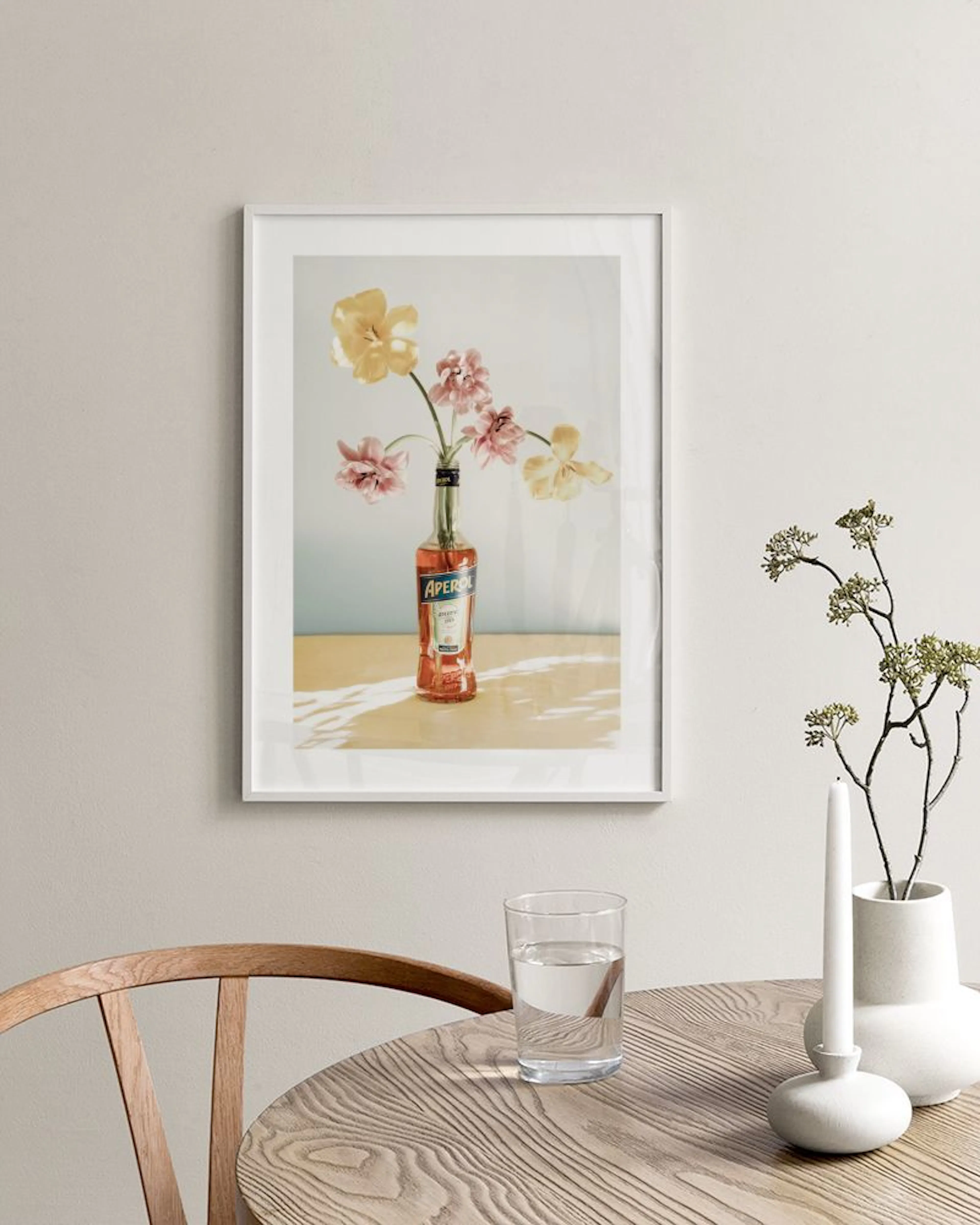 A print of yellow and pink flowers in an Aperol bottle, hanging above a white console table with decorative vases and books.