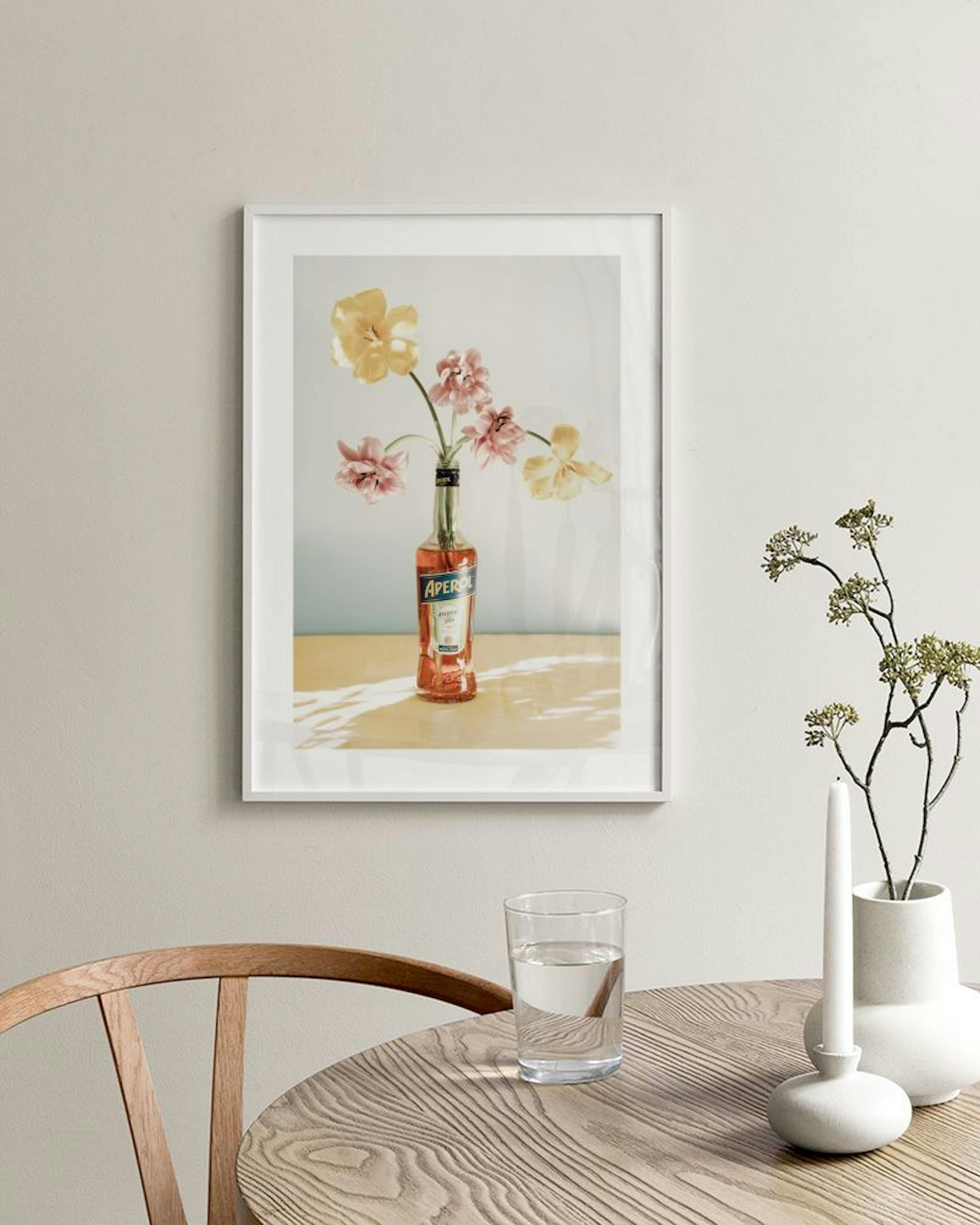 Poster van gele en roze bloemen in een Aperol fles, hangend boven een wit dressoir met vazen.