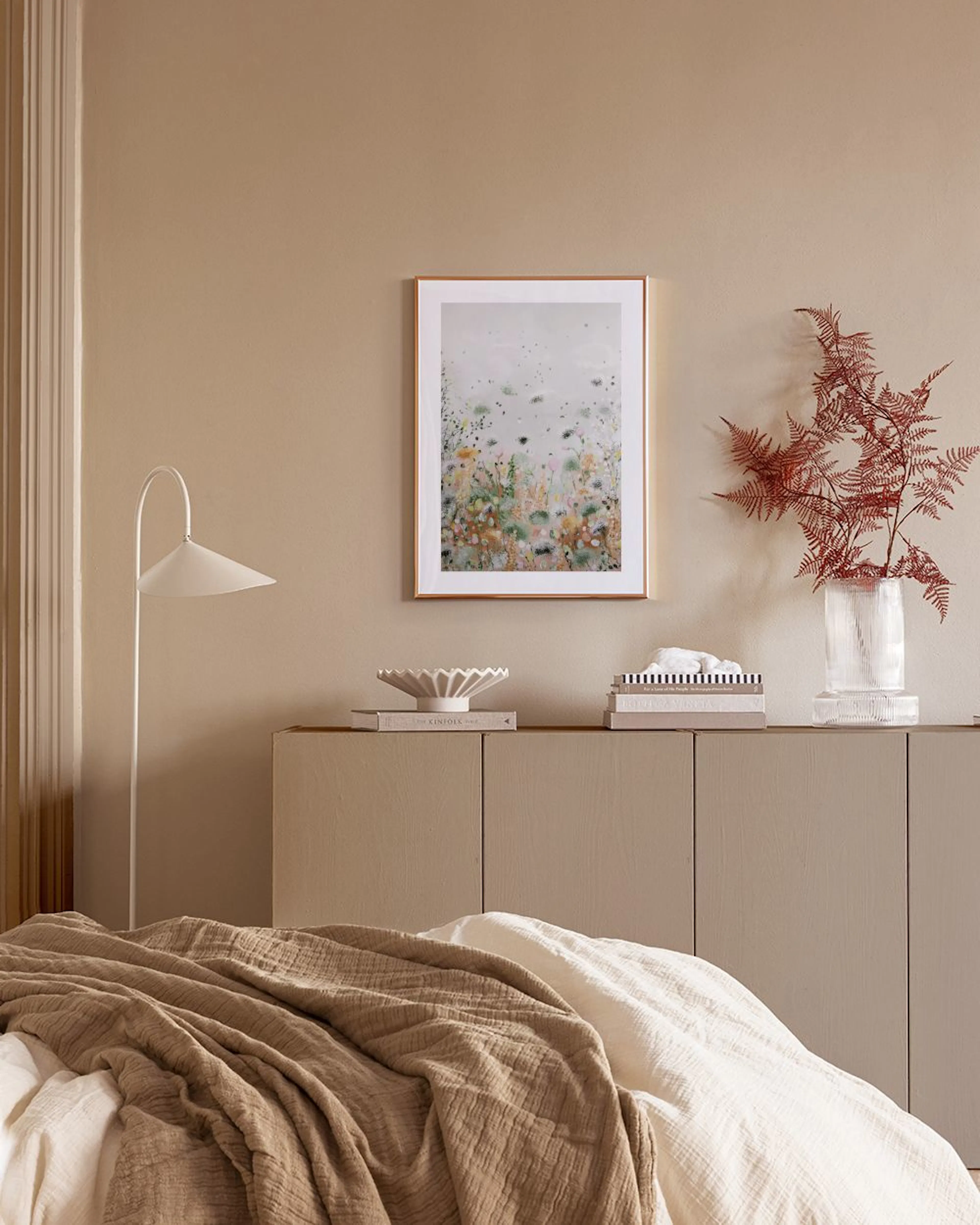 Een poster met een abstract bloemenveld in groen, oranje en roze tinten, hangend boven een wit dressoir met vazen en boeken.