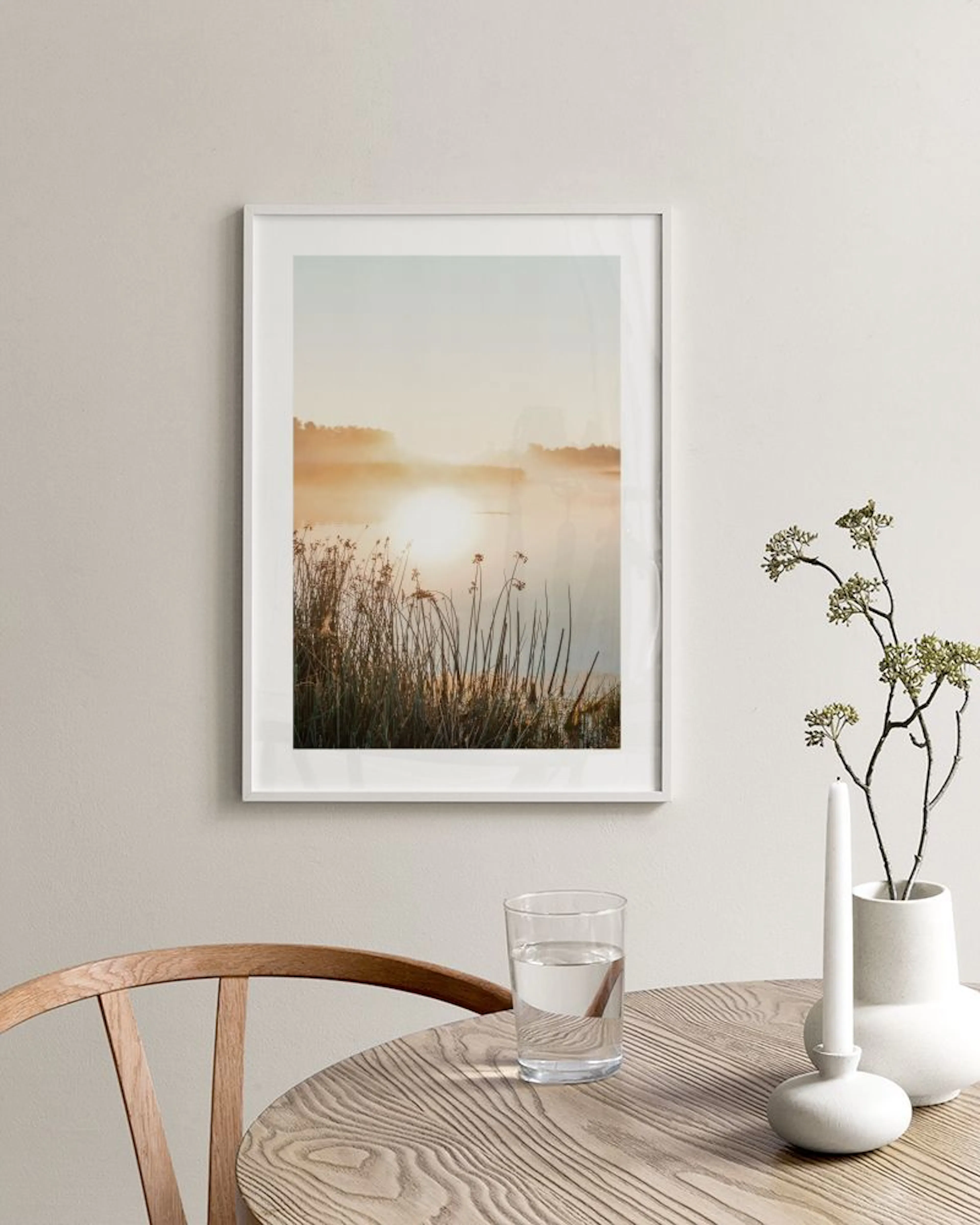 Poster eines nebligen Sonnenaufgangs über einem See mit Schilf, hängend über einem weißen Sideboard mit Vasen.