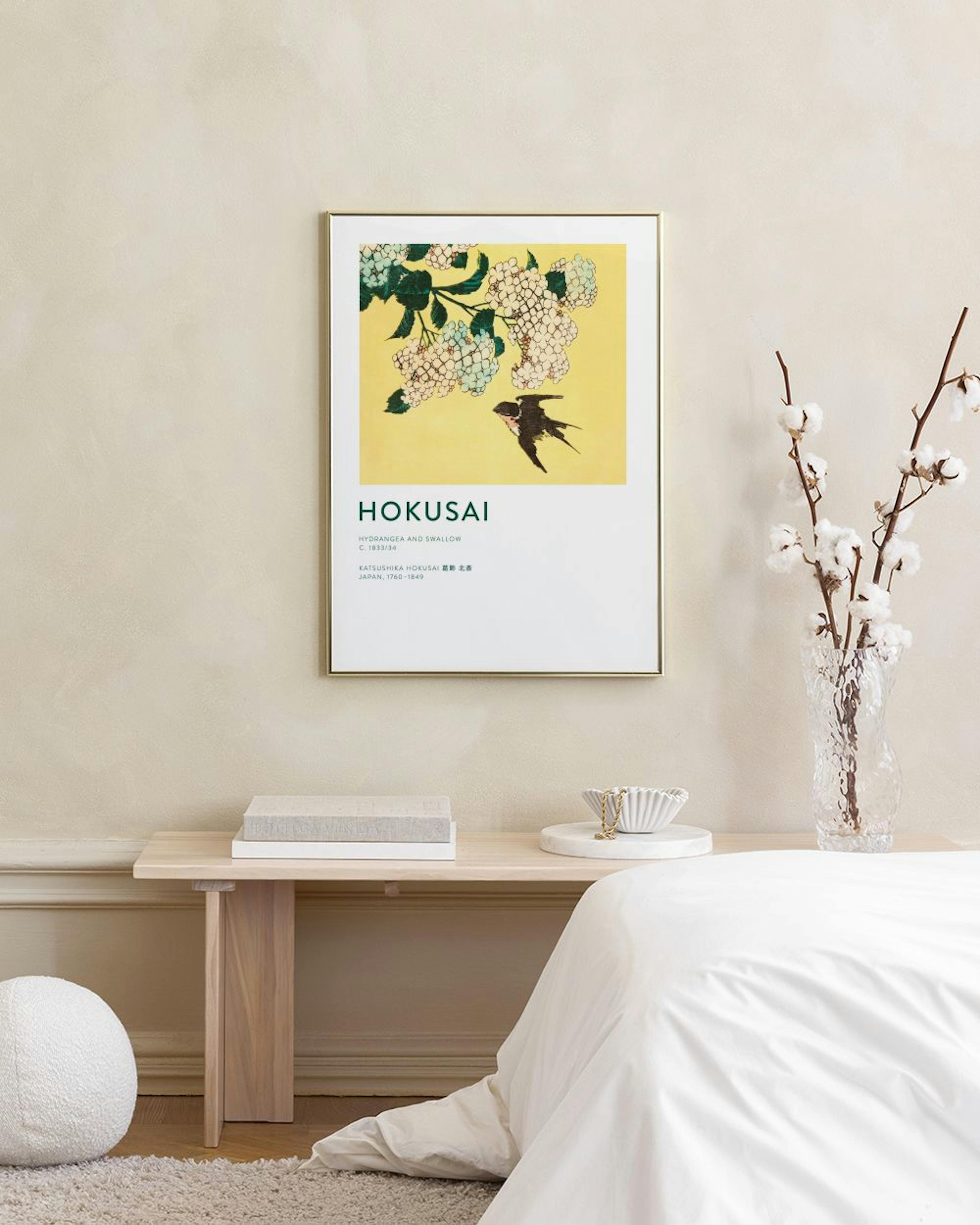 Póster de Hokusai con golondrina y flores blancas sobre fondo amarillo, colgando sobre un mueble blanco con un jarrón y un tabur