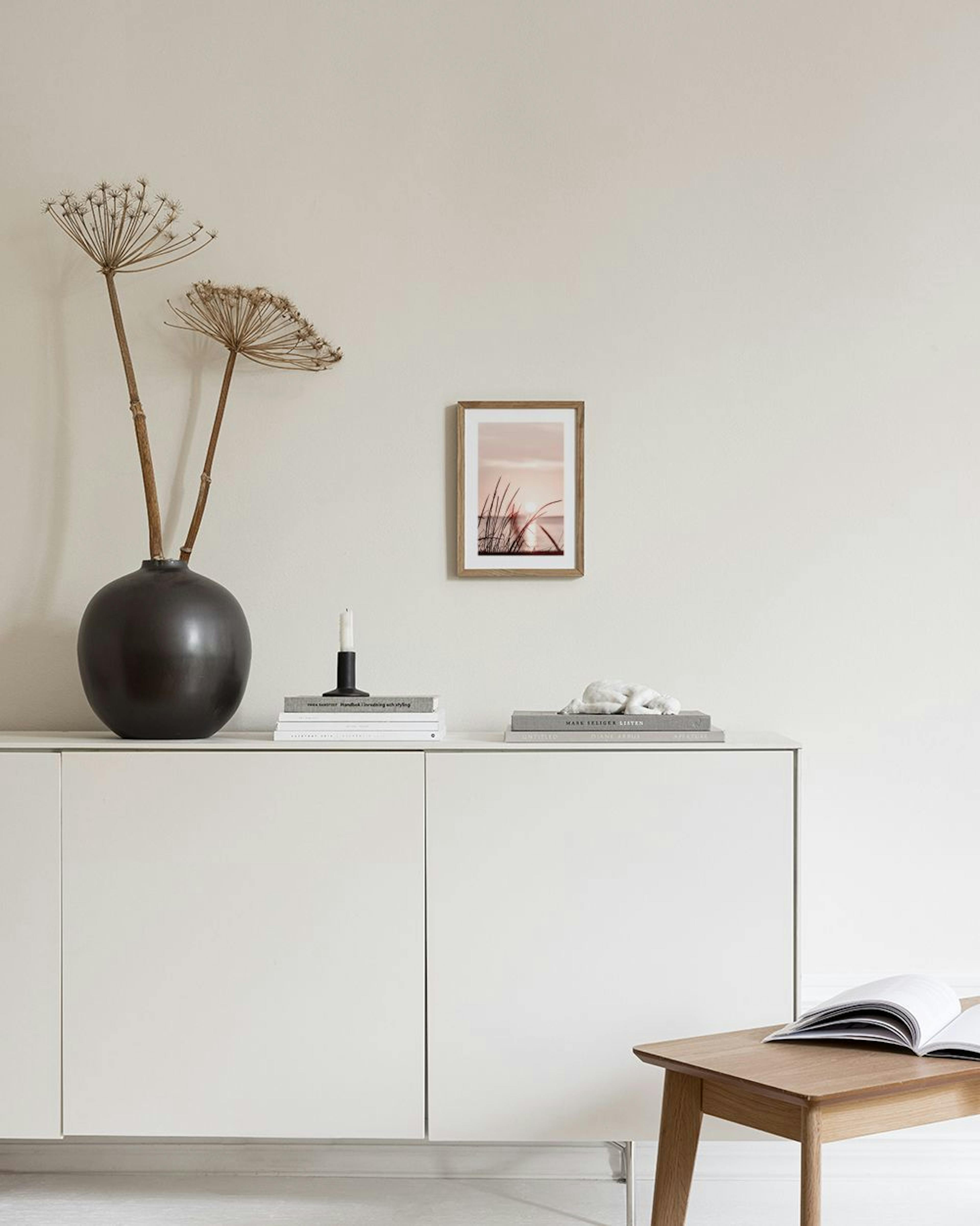 Poster van zonsondergang boven water met silhouetten van grassprieten, hangend boven een wit dressoir met vazen en boeken.