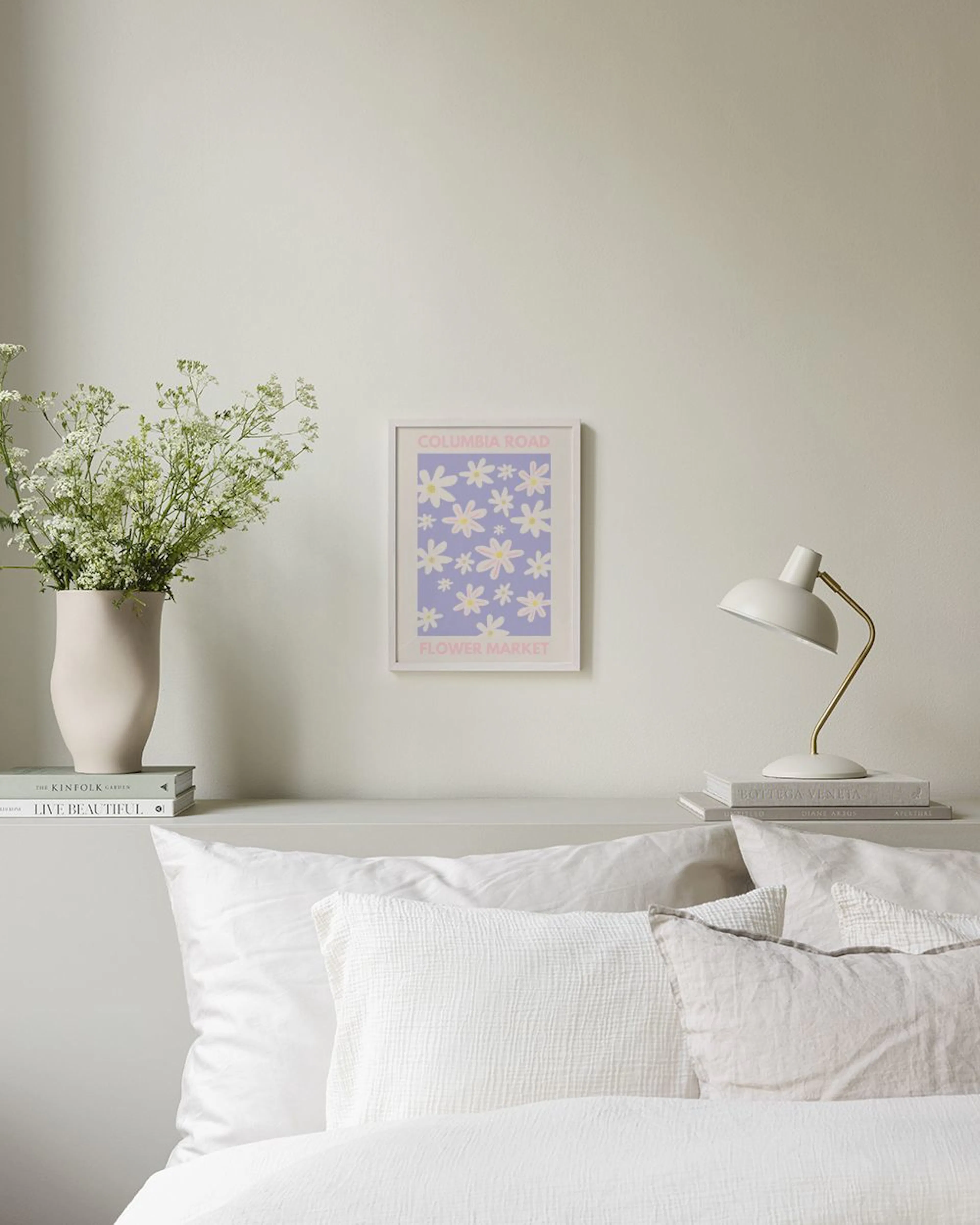 Póster Columbia Road Flower Market con diseño floral en tonos lila, blanco y rosa, colgado en una pared blanca sobre un taburete