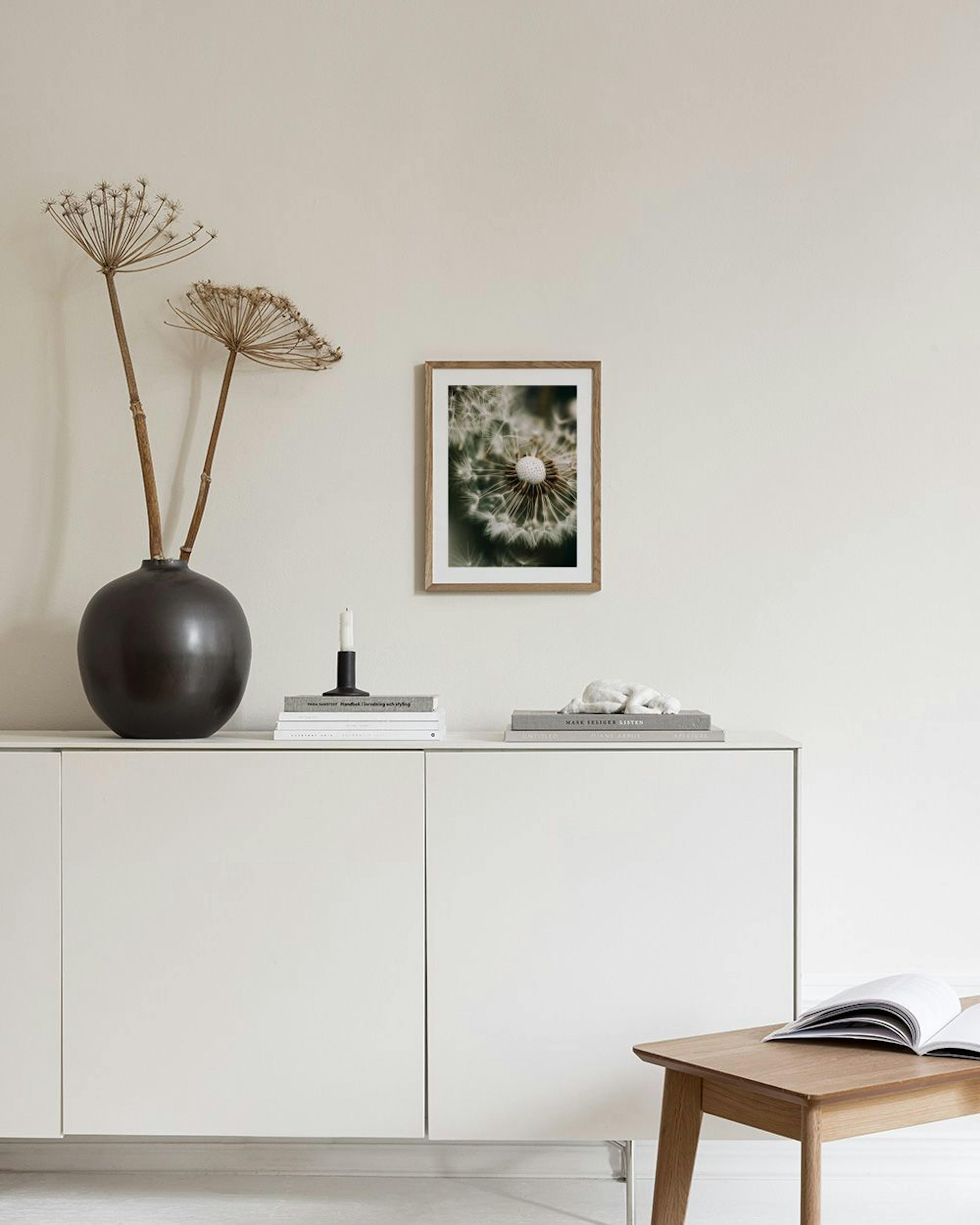 A print of a close-up dandelion seed head with white fluffy seeds, hanging above a white marble side table with two vases and bo