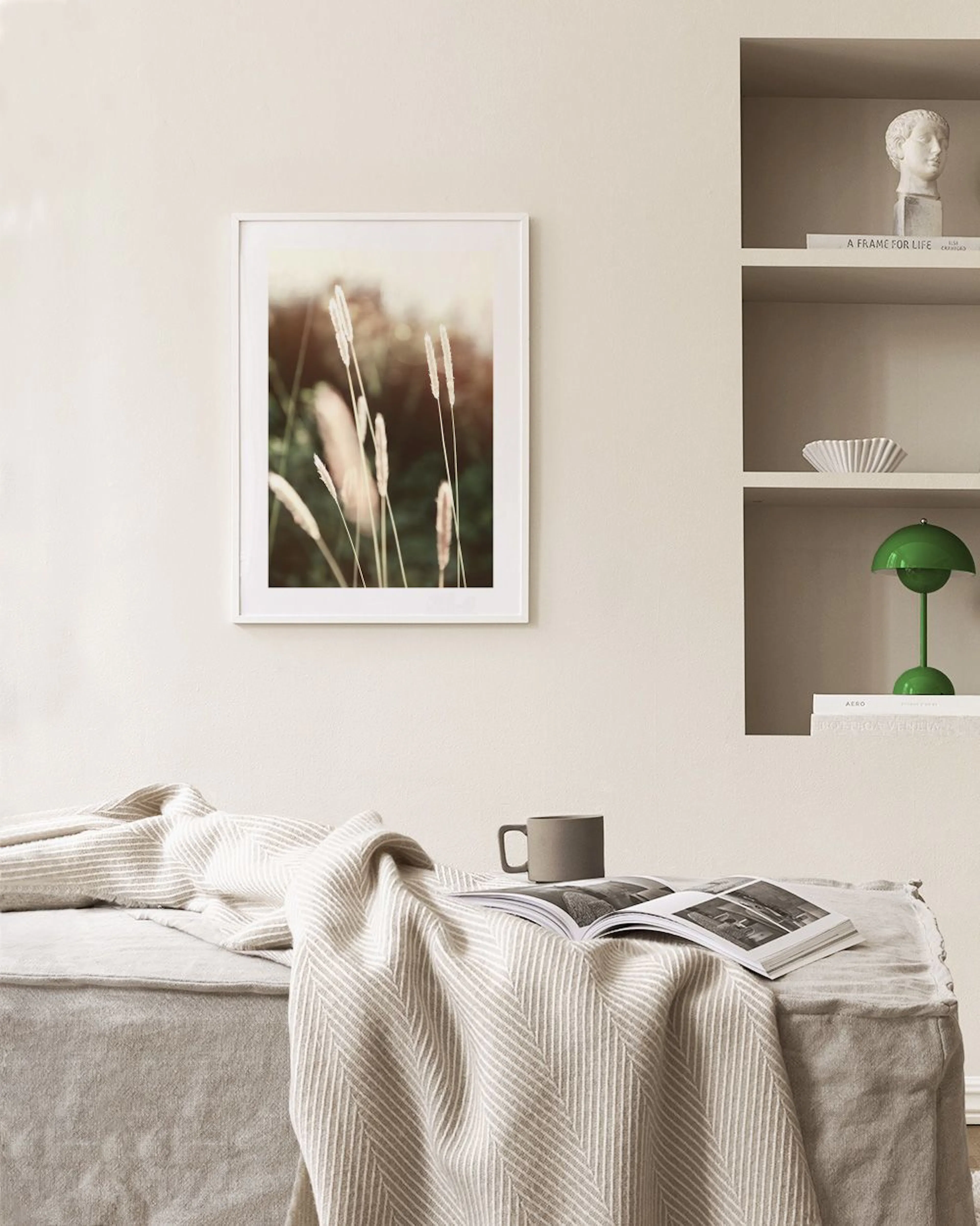 A nature print of light-coloured wild grasses against a soft, blurred green and brown background, hanging in a bright room with 