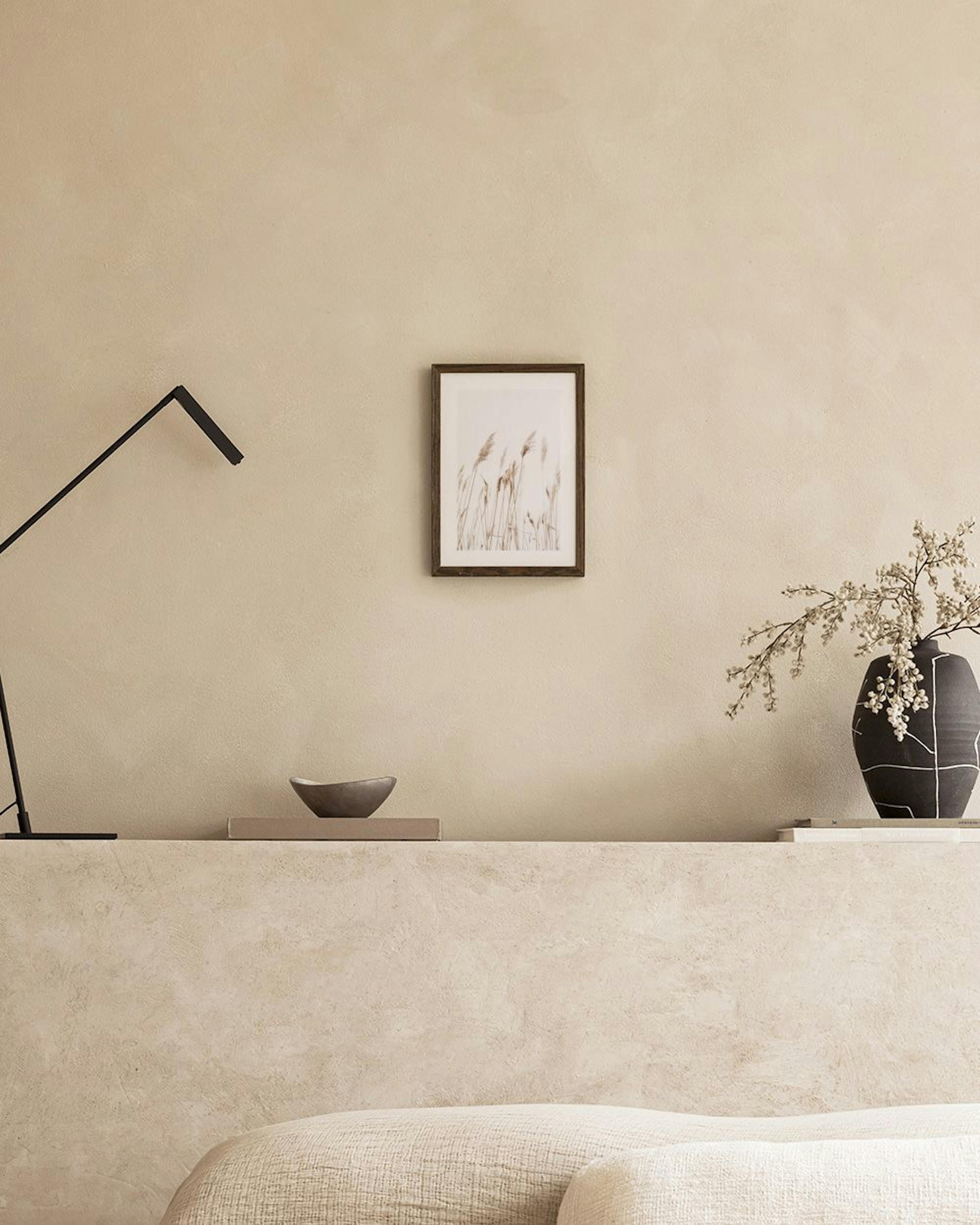 A print of tall, feathery dried reeds or grasses on a light background, hanging on a grey wall above a brown armchair.