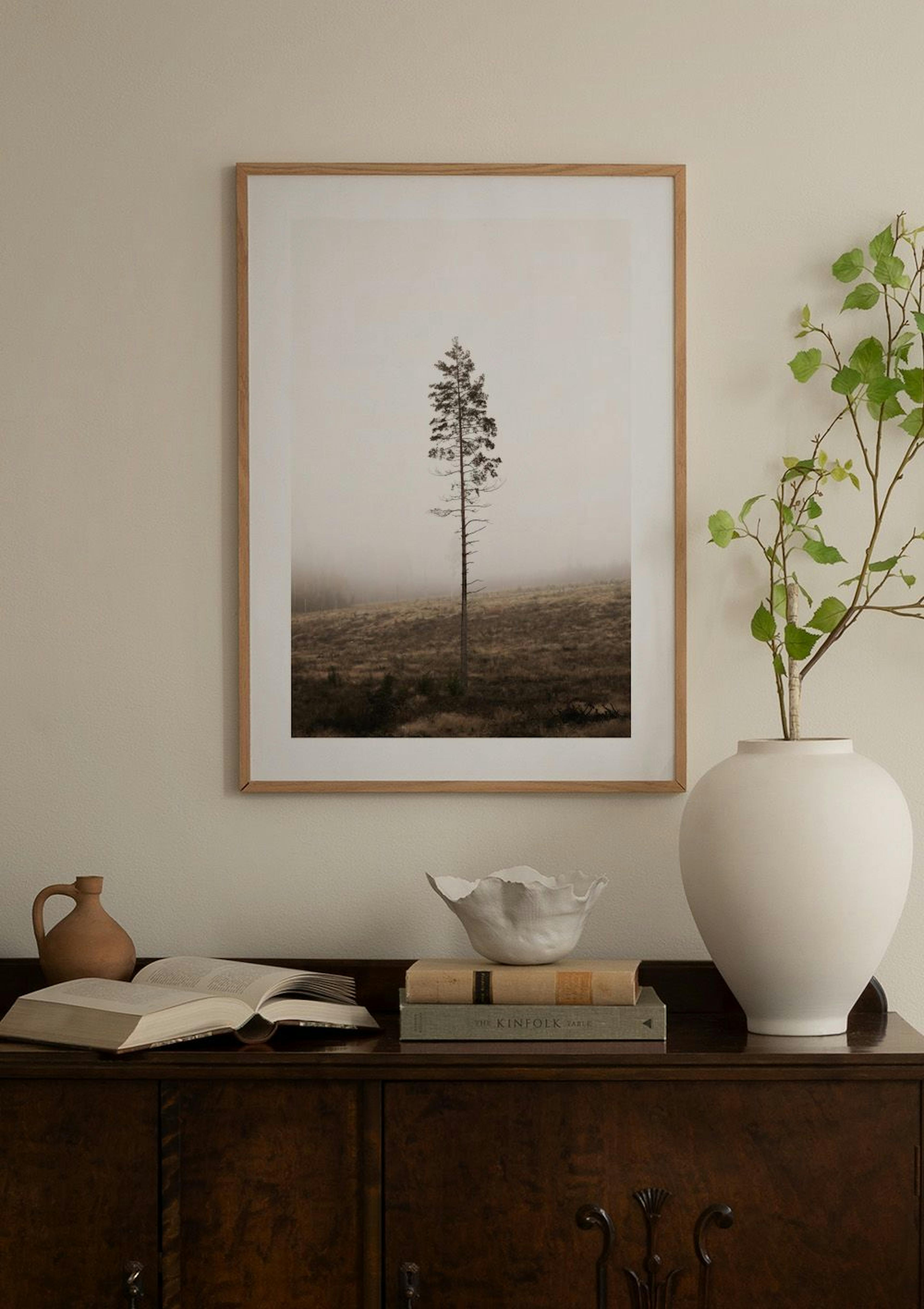 Mörk poster med ensamt träd i dimmigt landskap, hängande på en grå vägg ovanför ett vitt marmorbord.