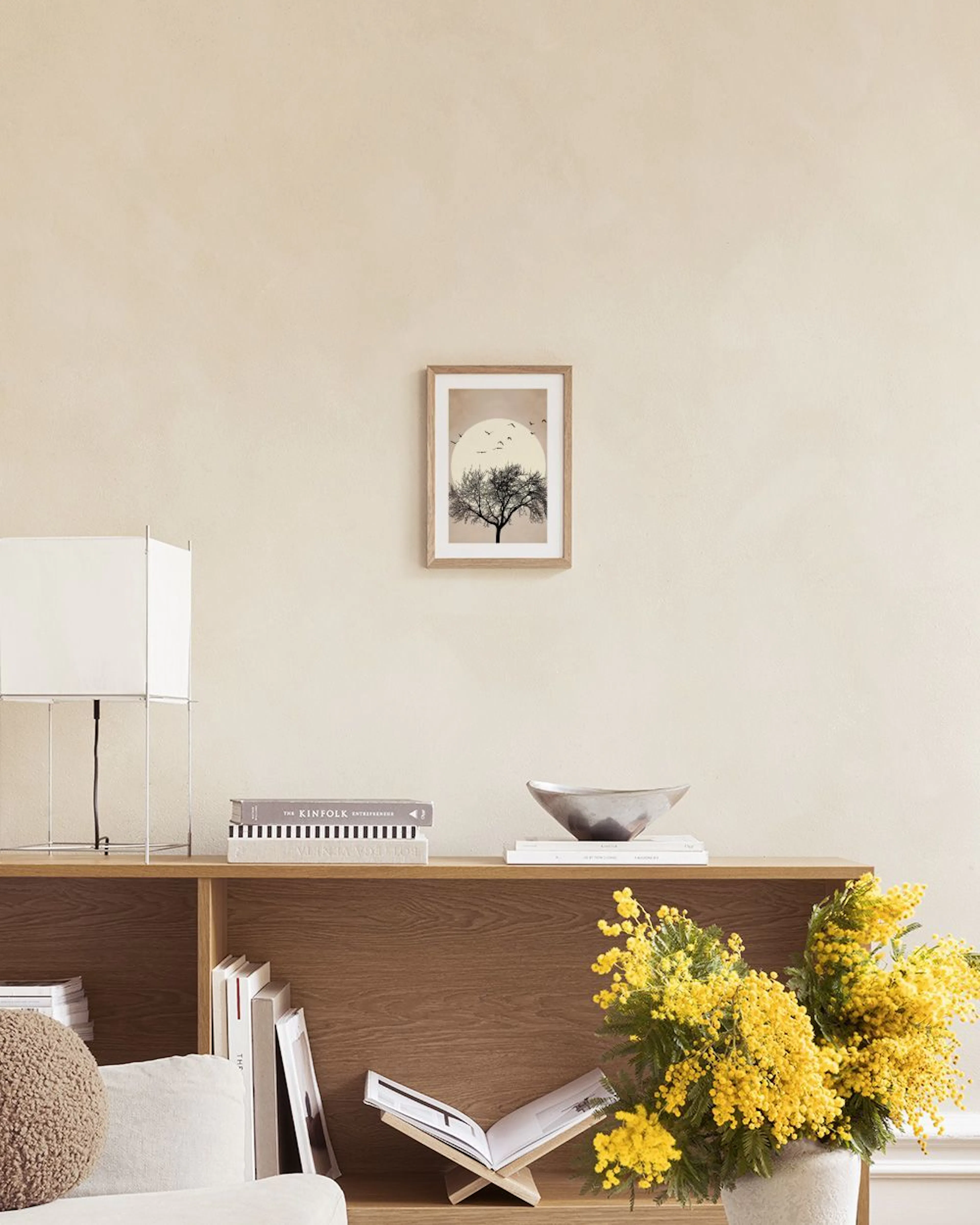 Poster van een kale boom silhouet voor een grote crème cirkel met vogels, hangend boven een beige fauteuil.