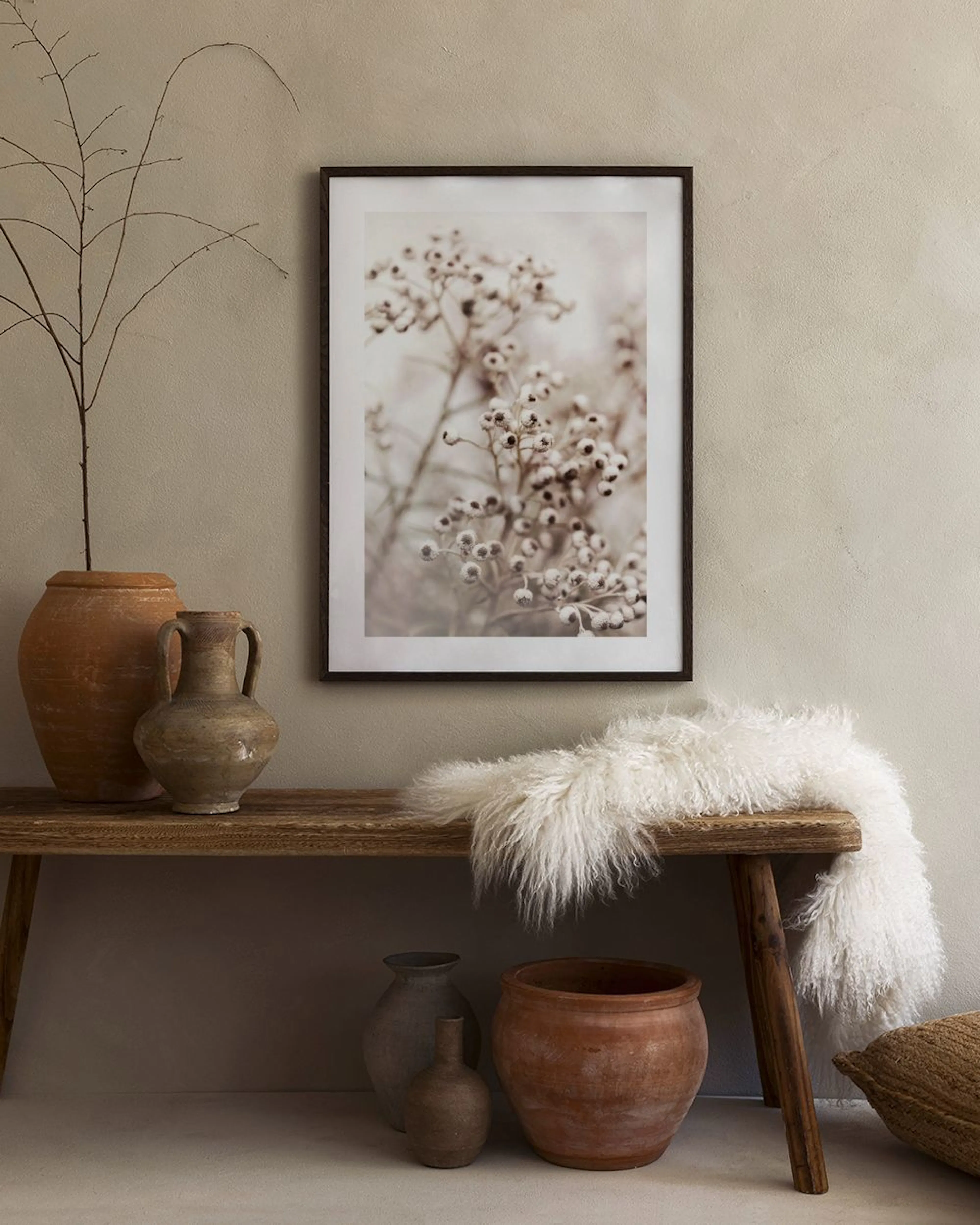 A print of dried white berries on a branch, hanging above a white marble side table with two white vases and two books.