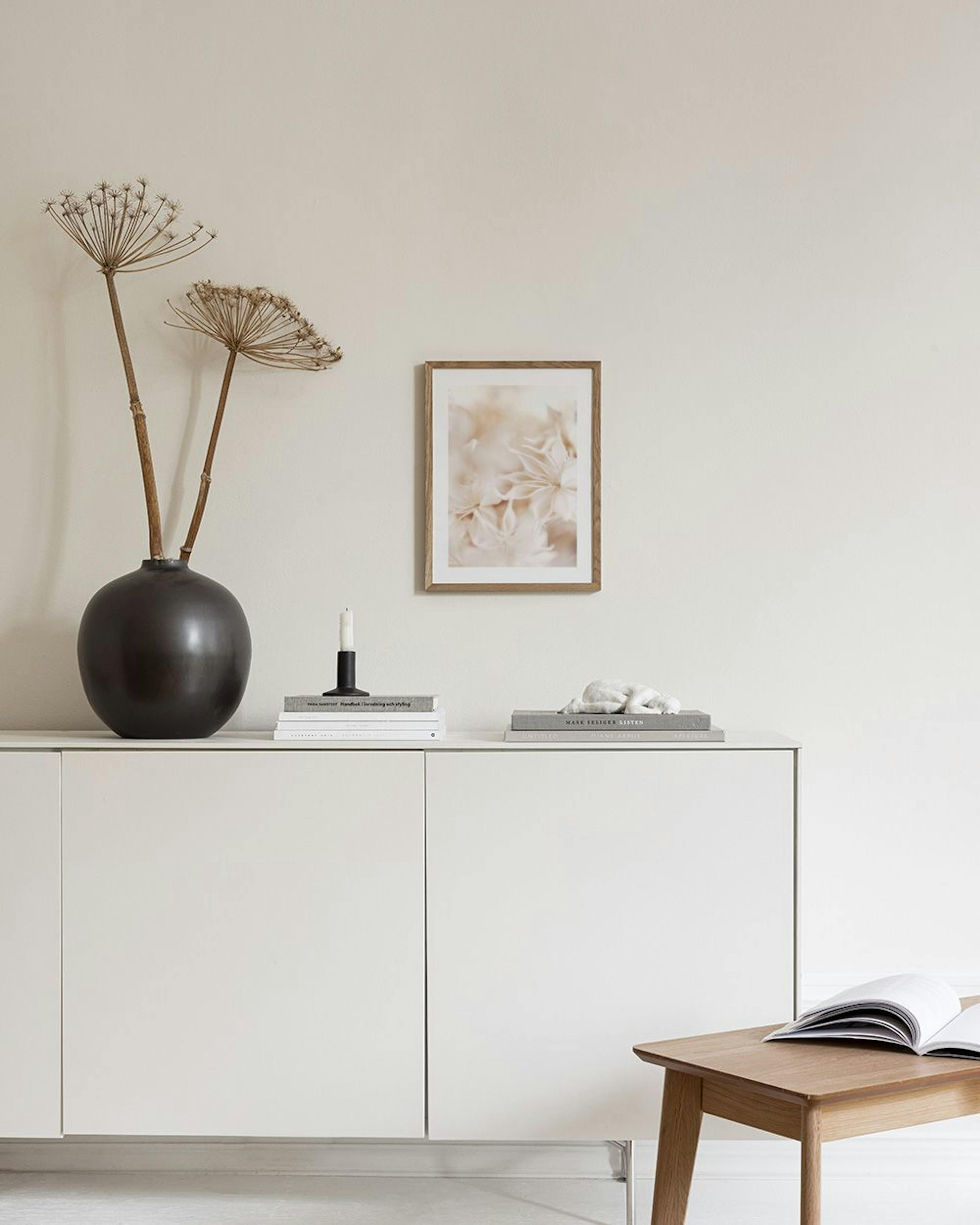 A print of soft, light pink flowers with delicate petals, hanging above a white marble side table with two white vases and stack