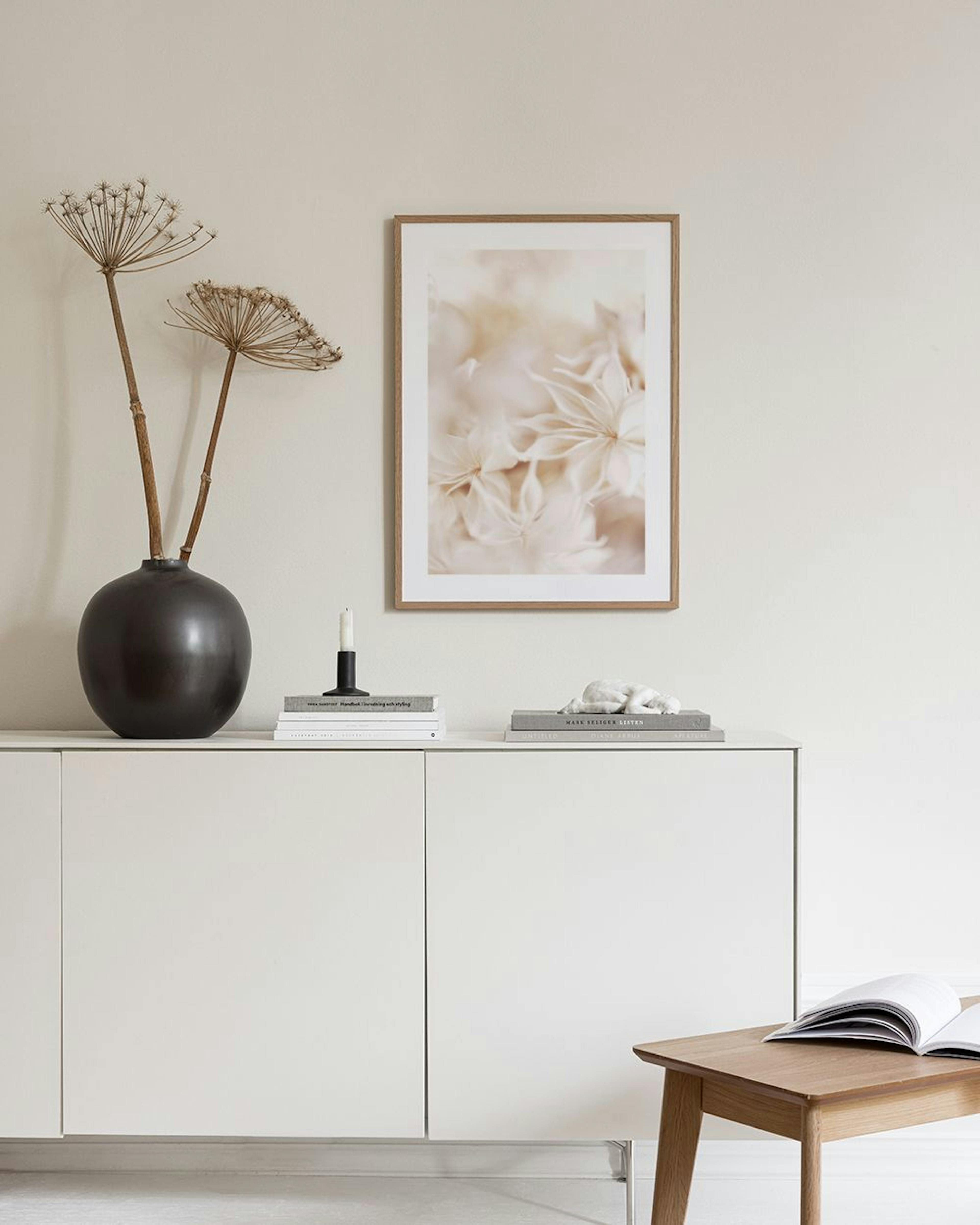 Plakat med lyse, bløde blomster i varme nuancer, hængende på en beige væg over et sidebord med bøger og vaser.