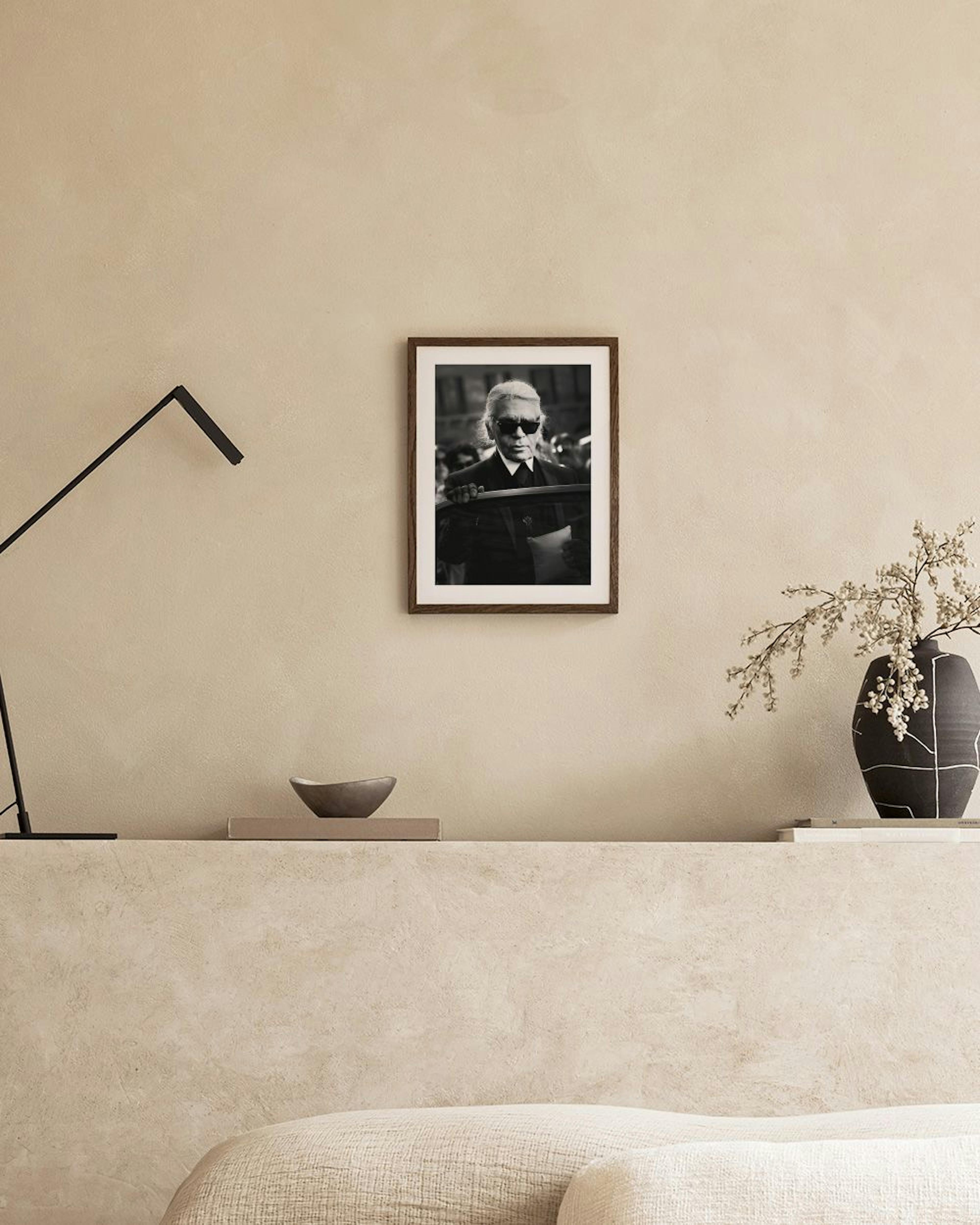 Póster en blanco y negro de Karl Lagerfeld con gafas de sol oscuras, colgado sobre un mueble blanco con un jarrón y un taburete.