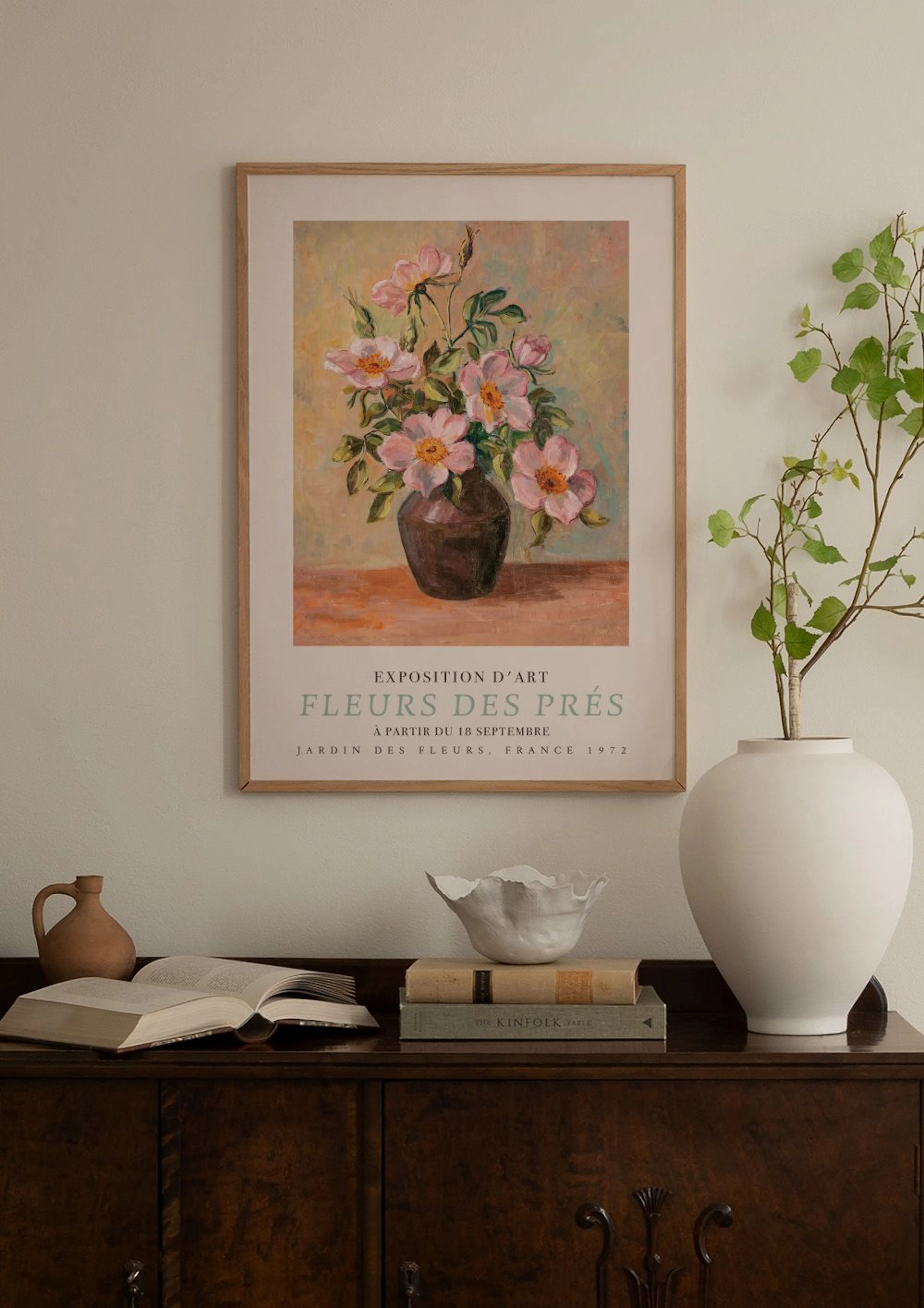 Plakat med maleri av rosa blomster i vase, henger på en lyserosa vegg over et hvitt skap med vaser og bøker.