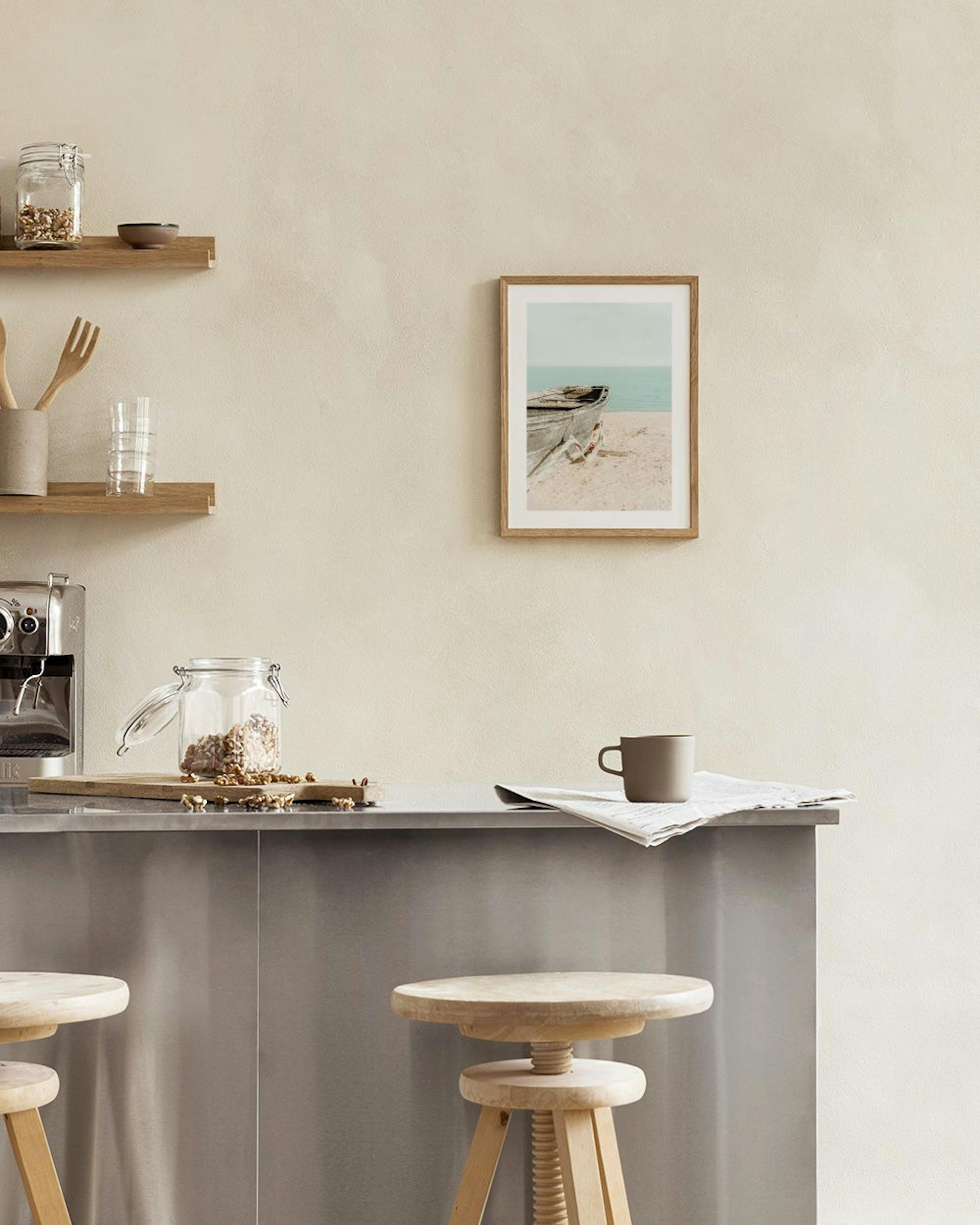 A print of an old wooden boat on a sandy beach next to calm blue water, hanging above a blue velvet chair in a minimalist room.