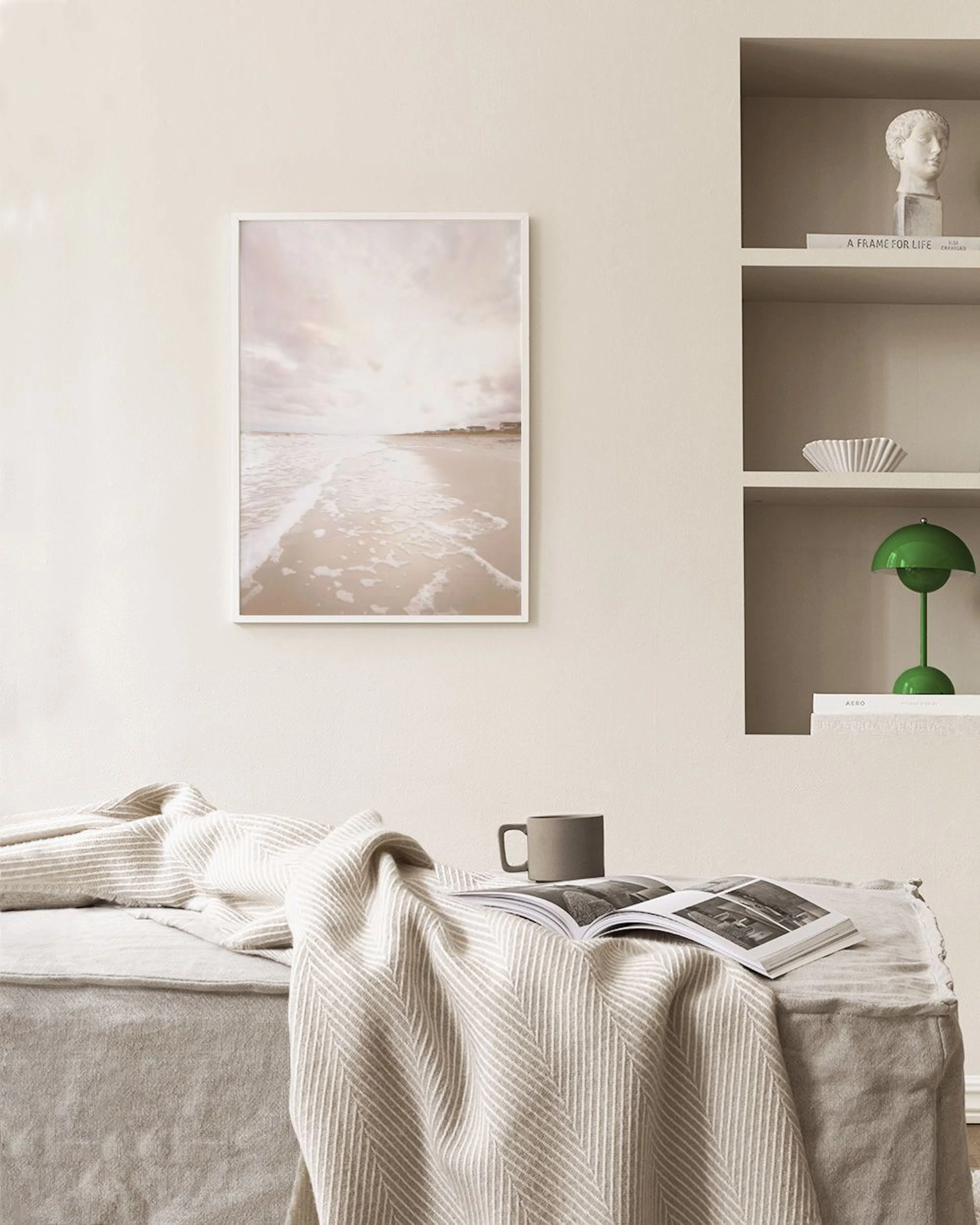 A print of a sandy beach and ocean waves under a cloudy sky, displayed above a white console table with three vases and two book