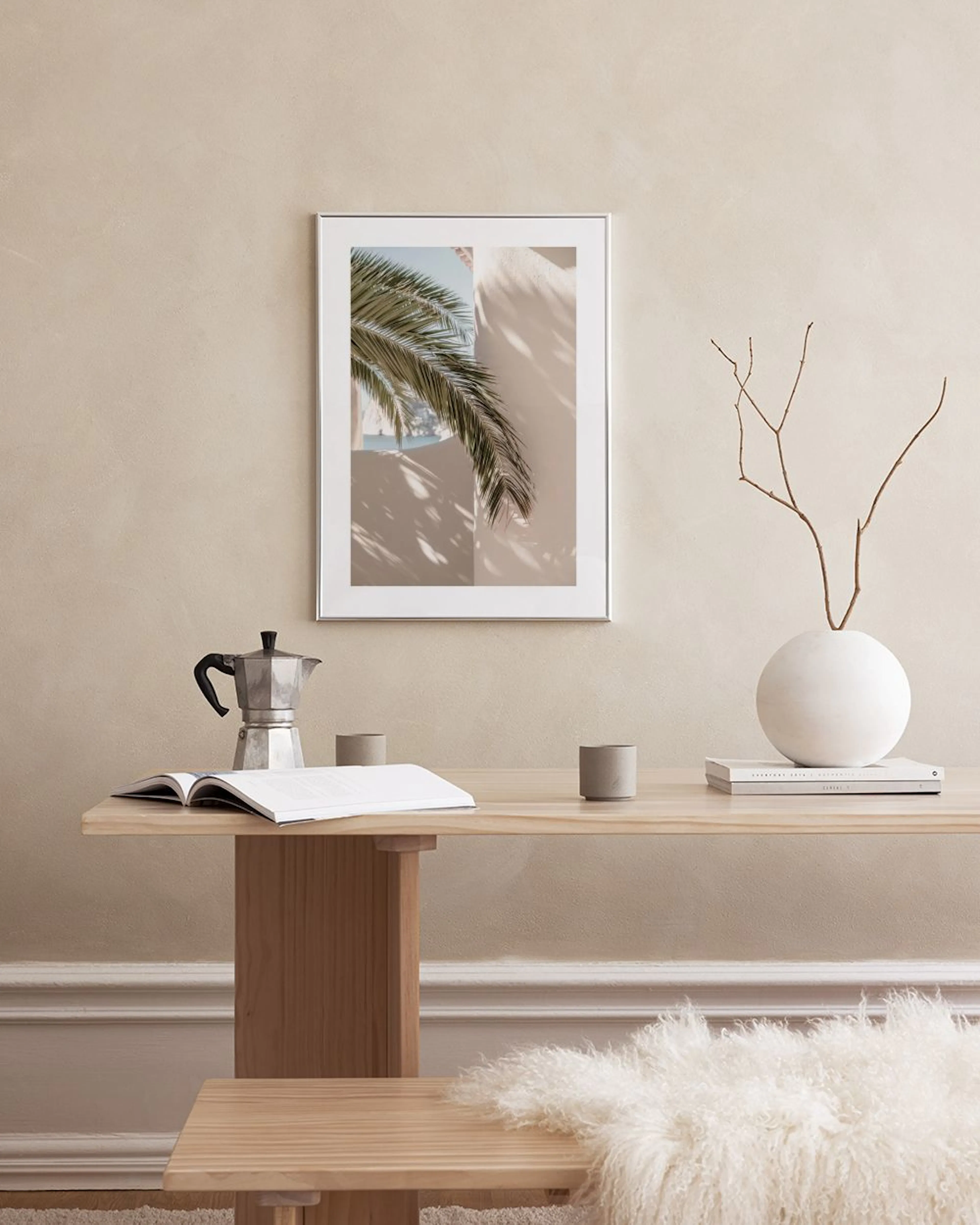 A tropical print of a palm tree against a beige wall, hanging above a white marble table with two vases and books.