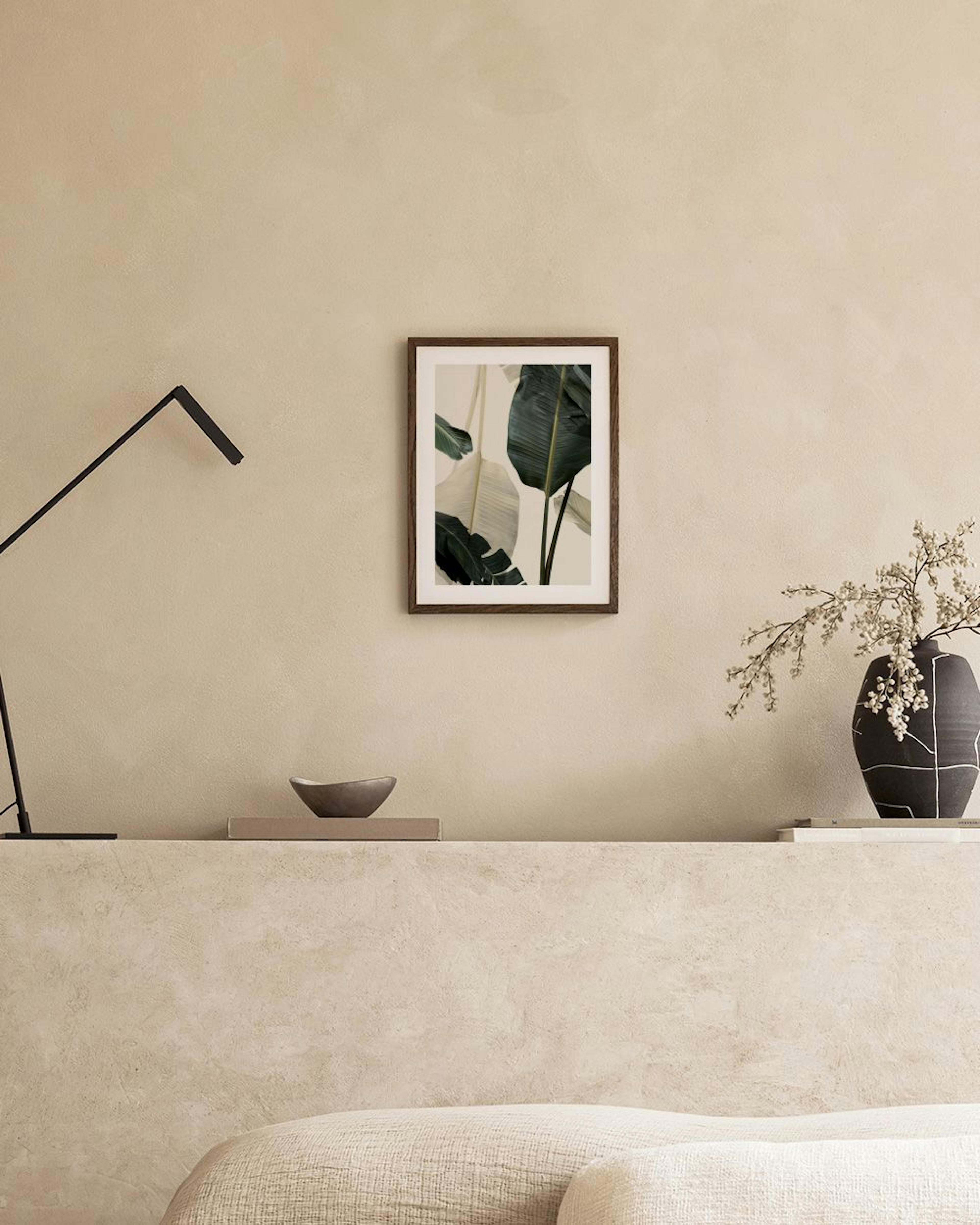 Póster de hojas de palma verde oscuro y beige claro, en una pared gris sobre un mueble blanco con libros, un jarrón y una silla.