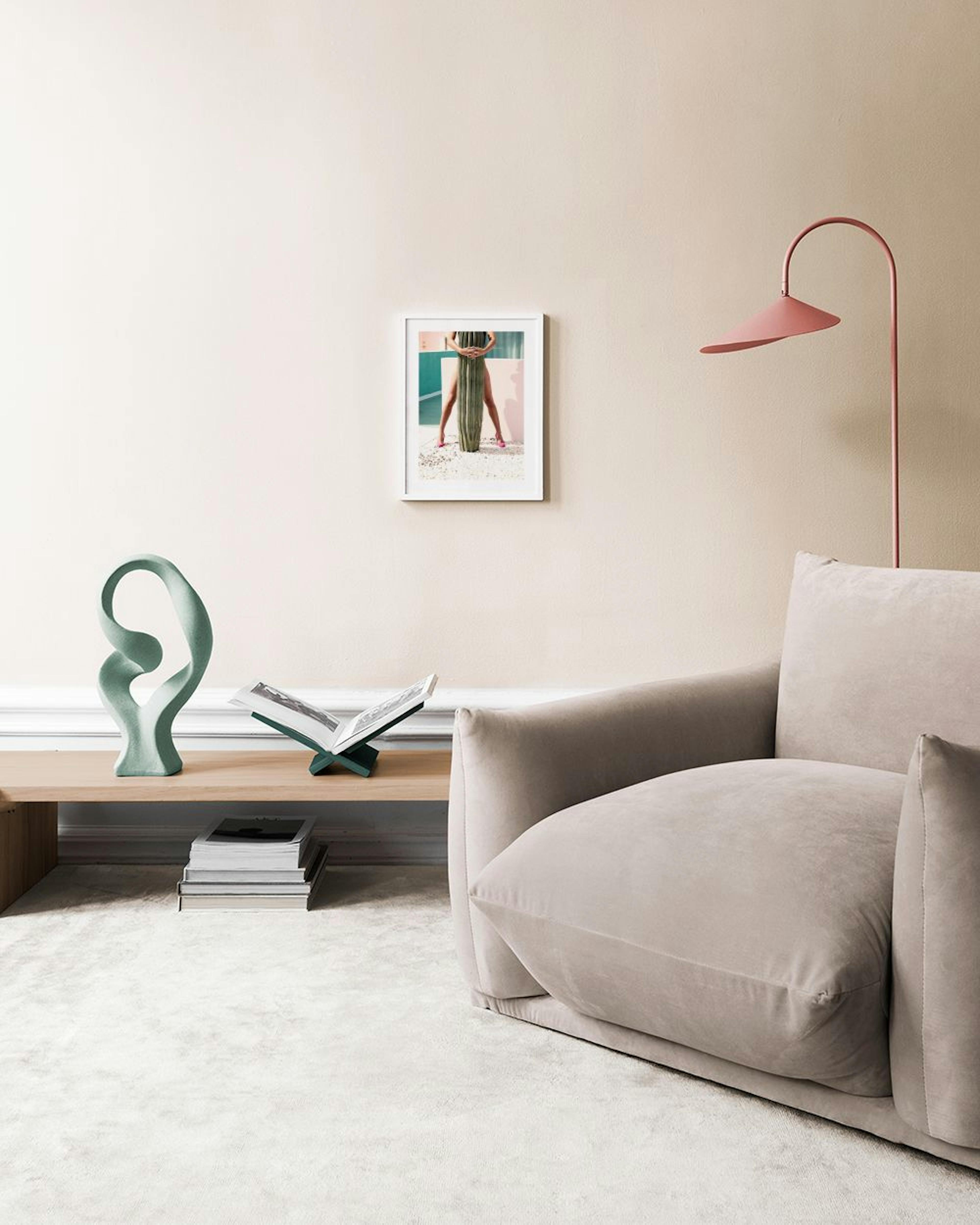 Poster de fotografia com pernas femininas à volta de um cato, em fundo rosa e verde, sobre móvel branco.