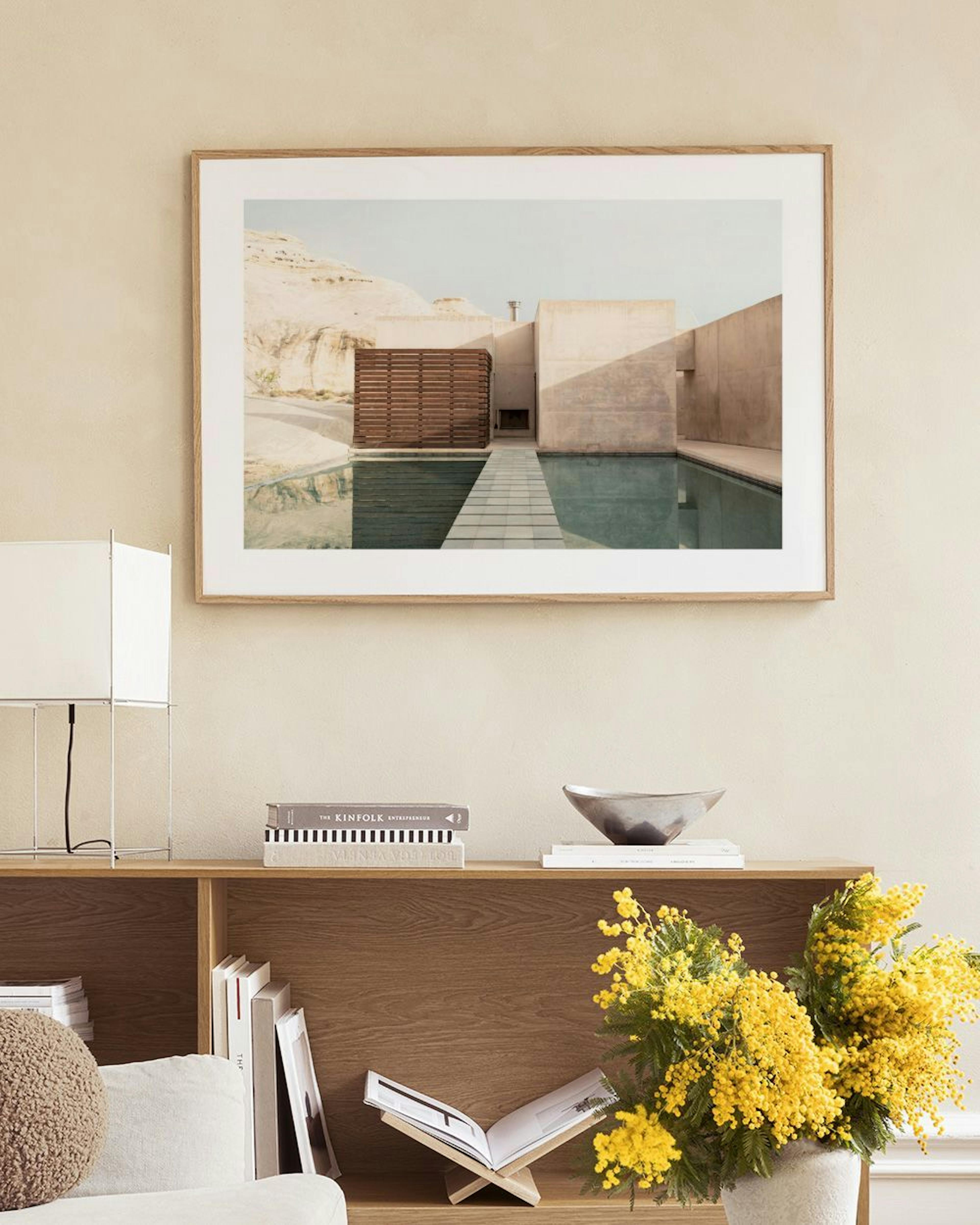 A print of a minimalist desert oasis building with a pool, hanging above a light wood stool with books and a terracotta vase.
