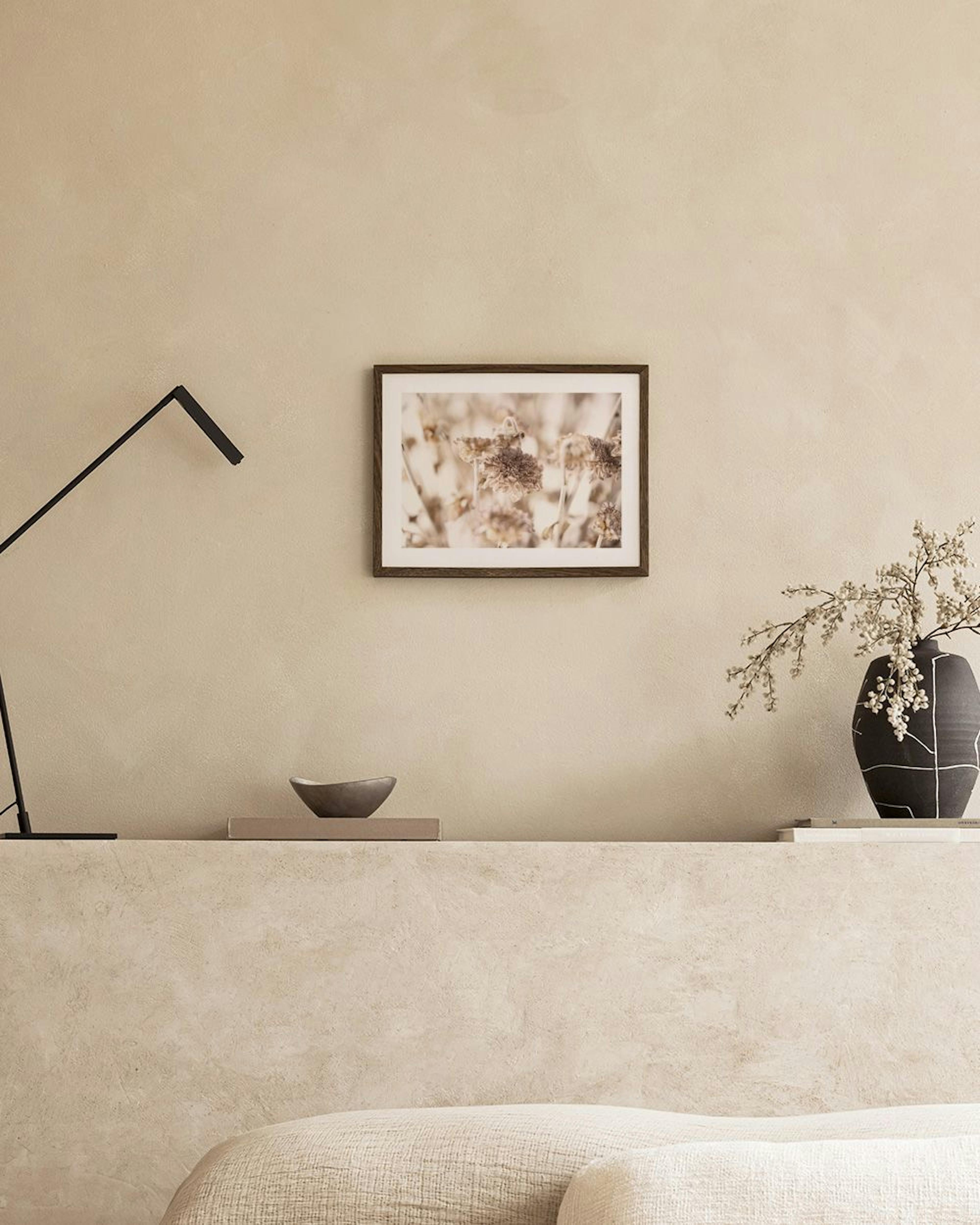 A print of dried flowers in sepia tones, hanging above a light wooden bench with books and a terracotta pot with pampas grass.