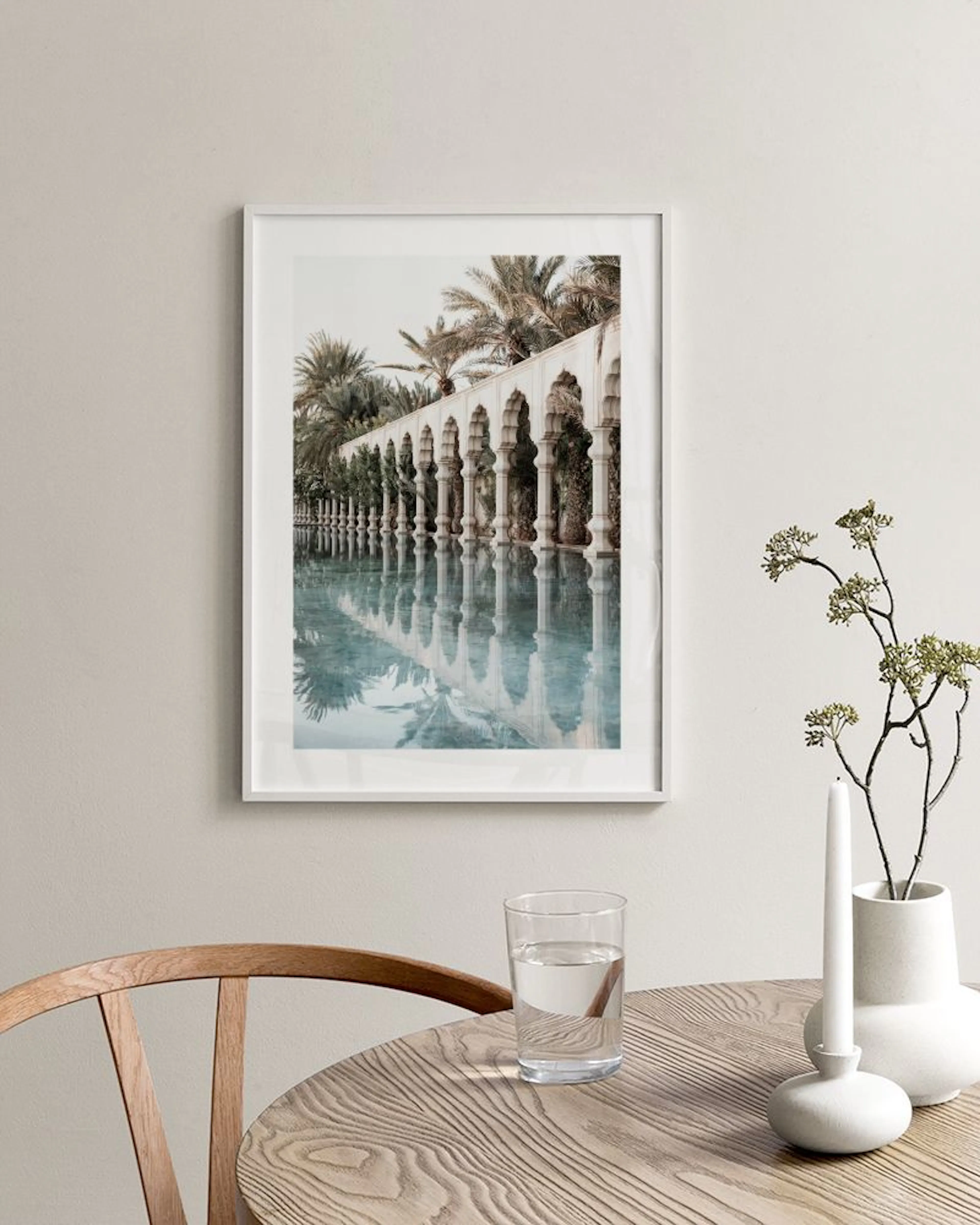 Architectural print with arches reflected in still water, surrounded by palm trees, hanging in a room with a wicker chair.