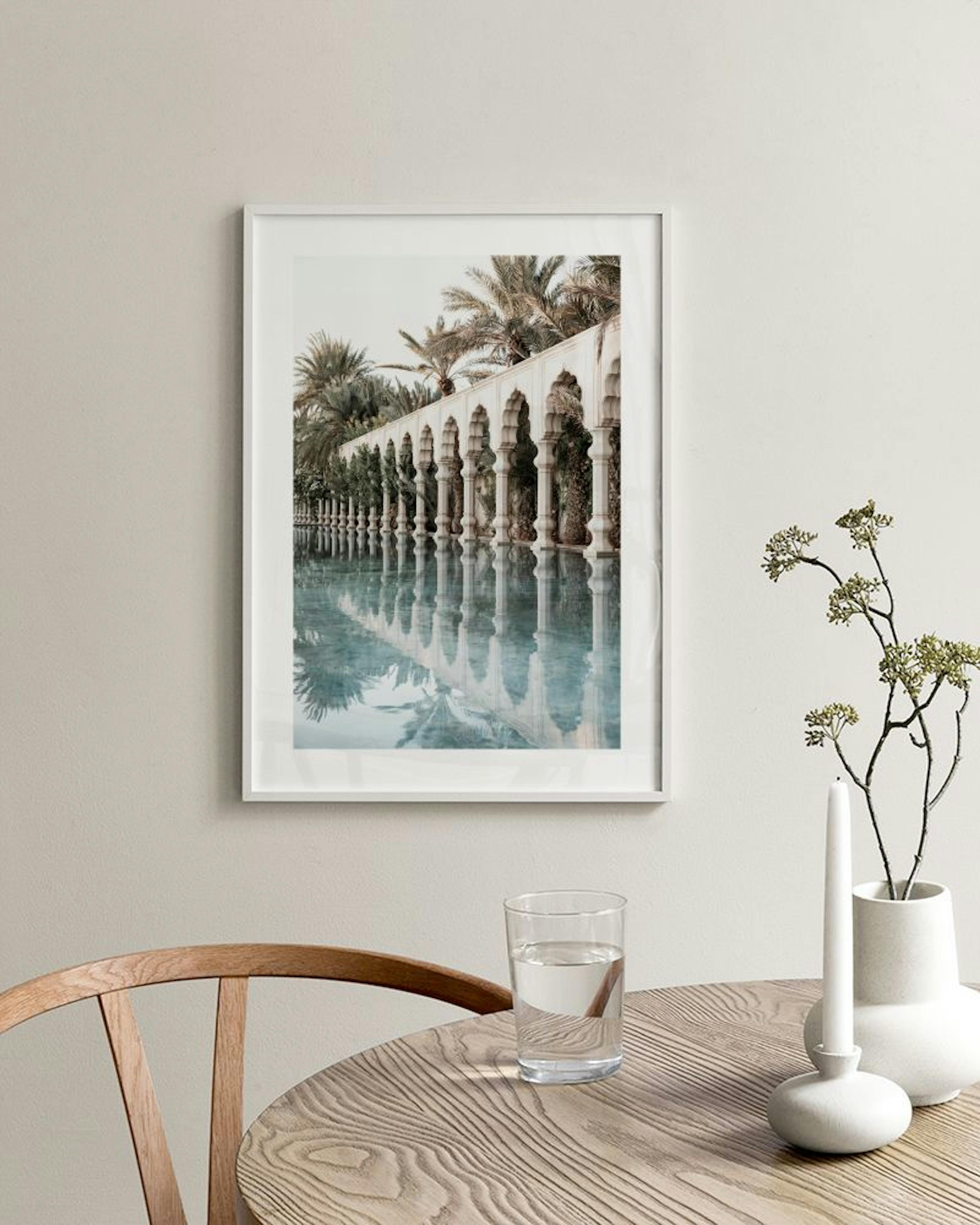 Poster van een witte zuilengalerij met palmbomen, weerspiegeld in lichtblauw water, hangend boven een rotan stoel.