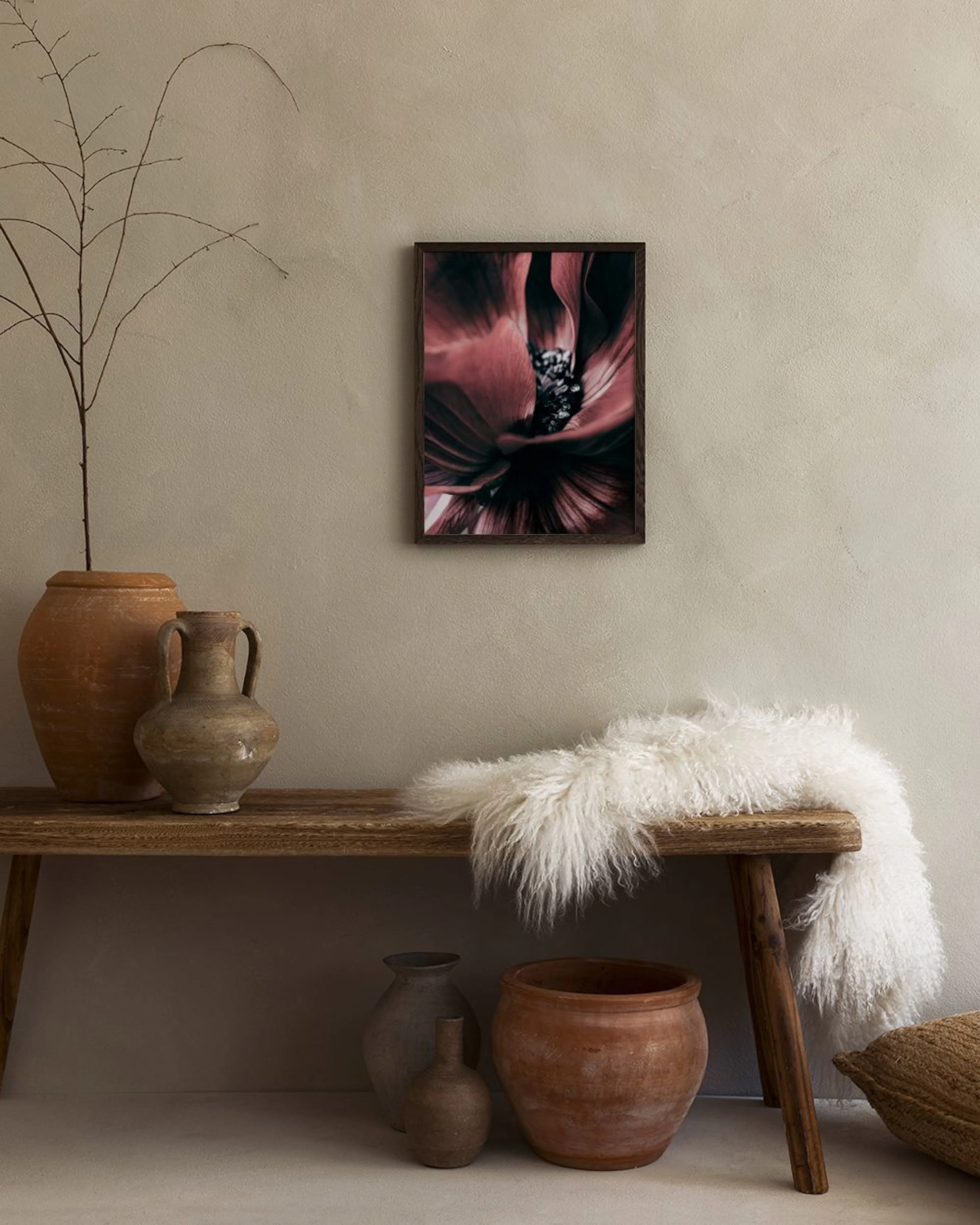 Poster med en mörkrosa pionblomma, hängande på en ljus vägg ovanför ett mörkbrunt sidobord med en byst och en puff.