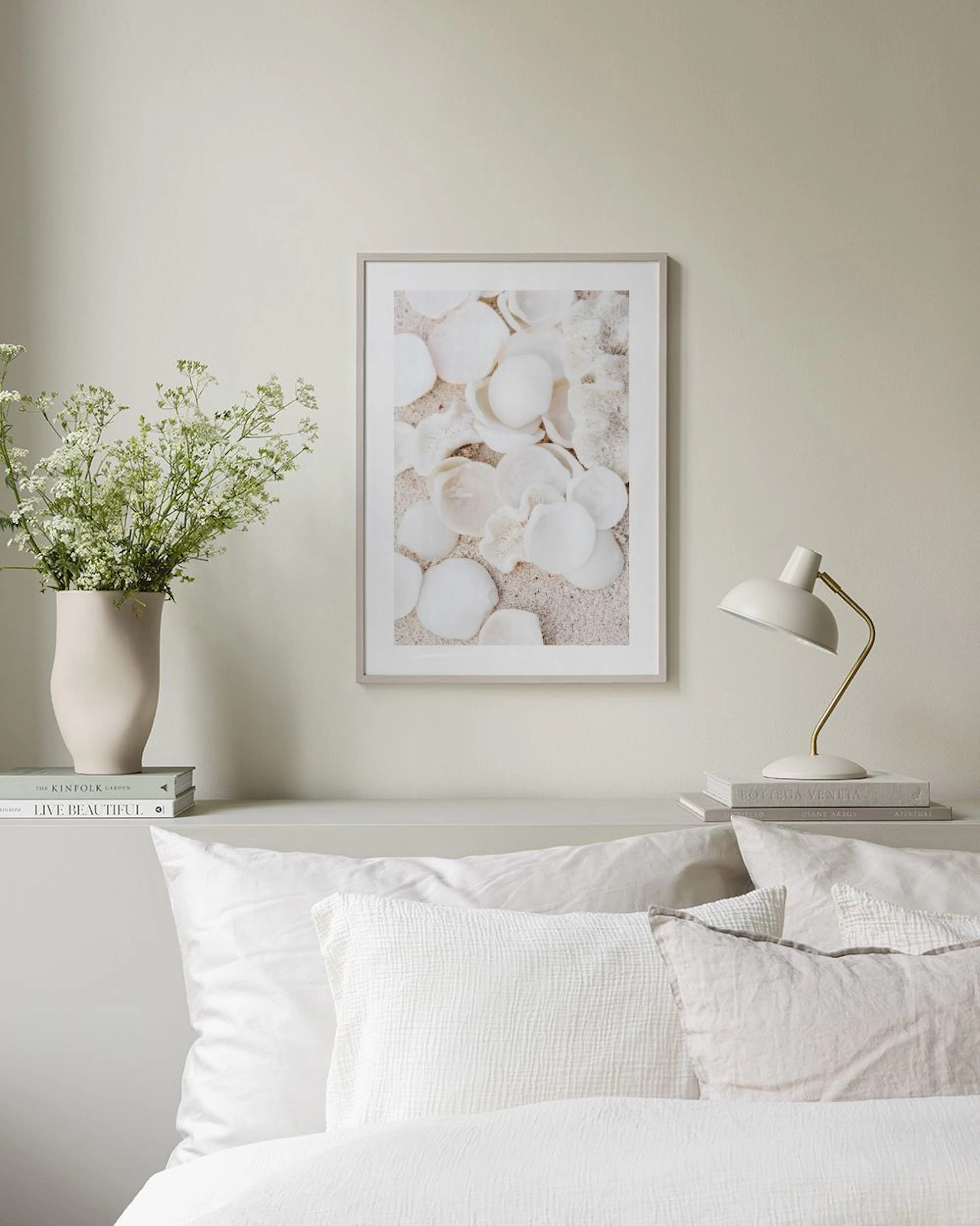 A print of white and cream seashells and coral on sand, hanging above a bedside table with books and a glass of water.