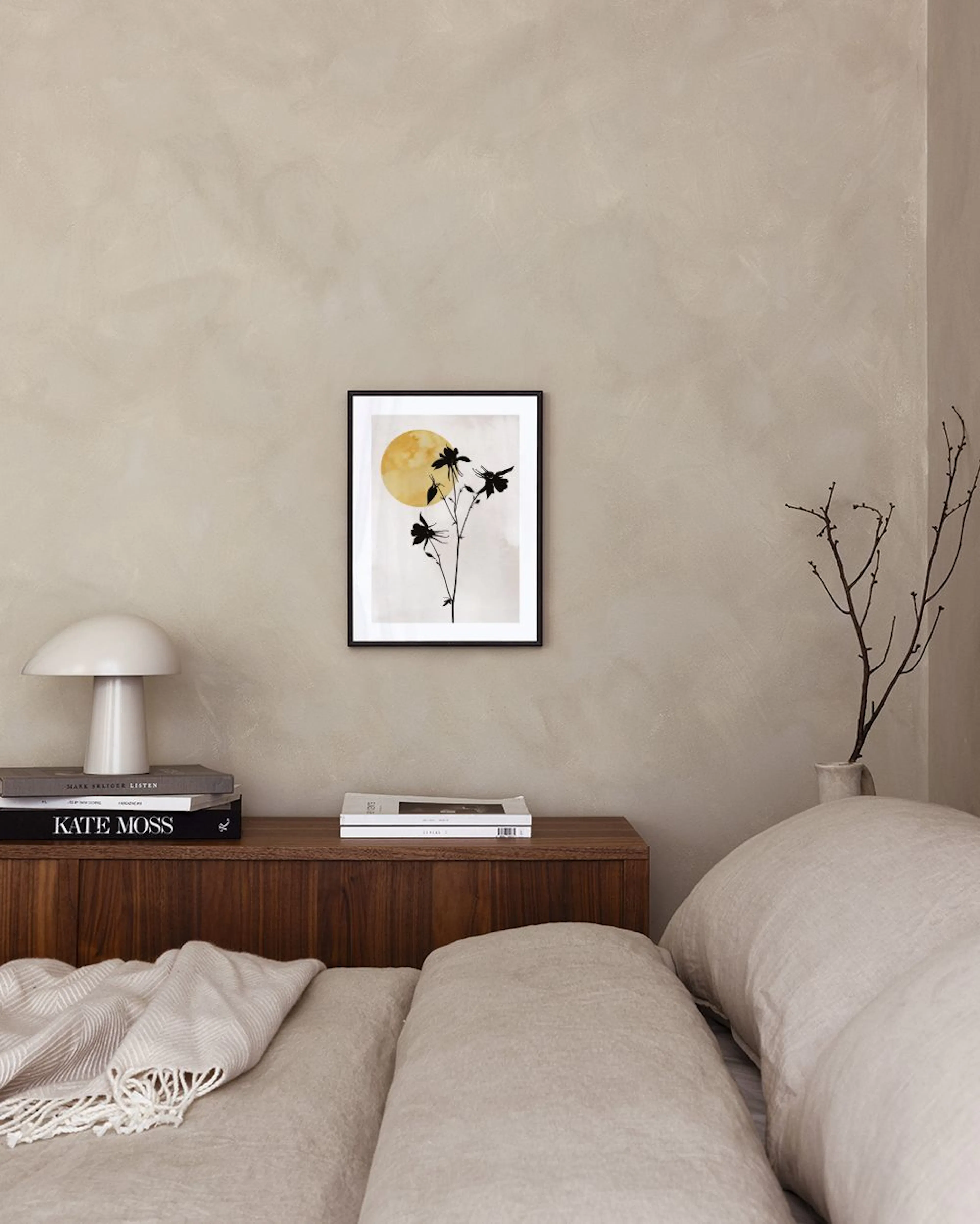 Poster med svarta silhuetter av blommor mot en gul sol på ljus bakgrund, hängande i ett rum med en brun fåtölj.