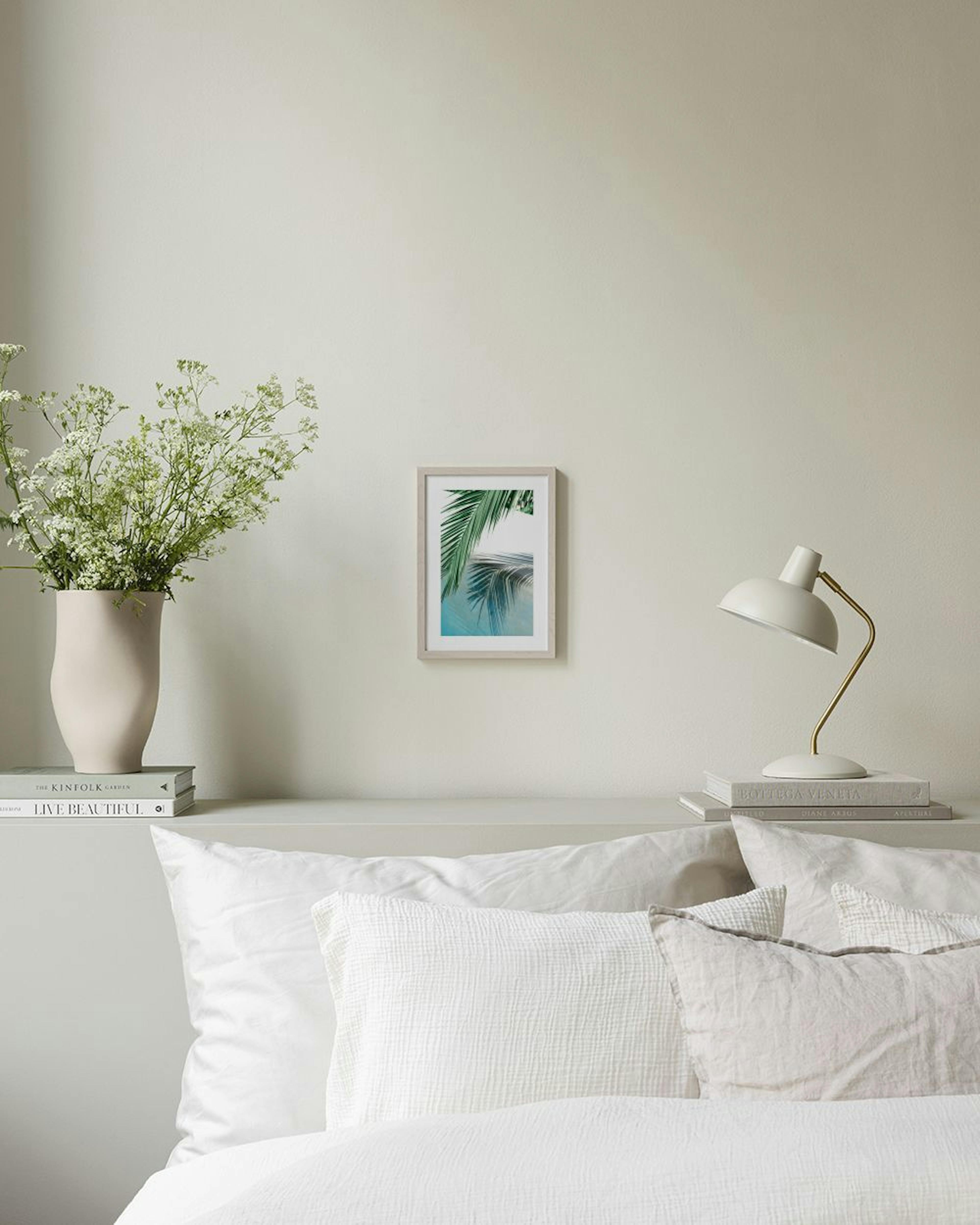 Print of a green palm leaf with its reflection on blue water, hanging above a woven bench with books.