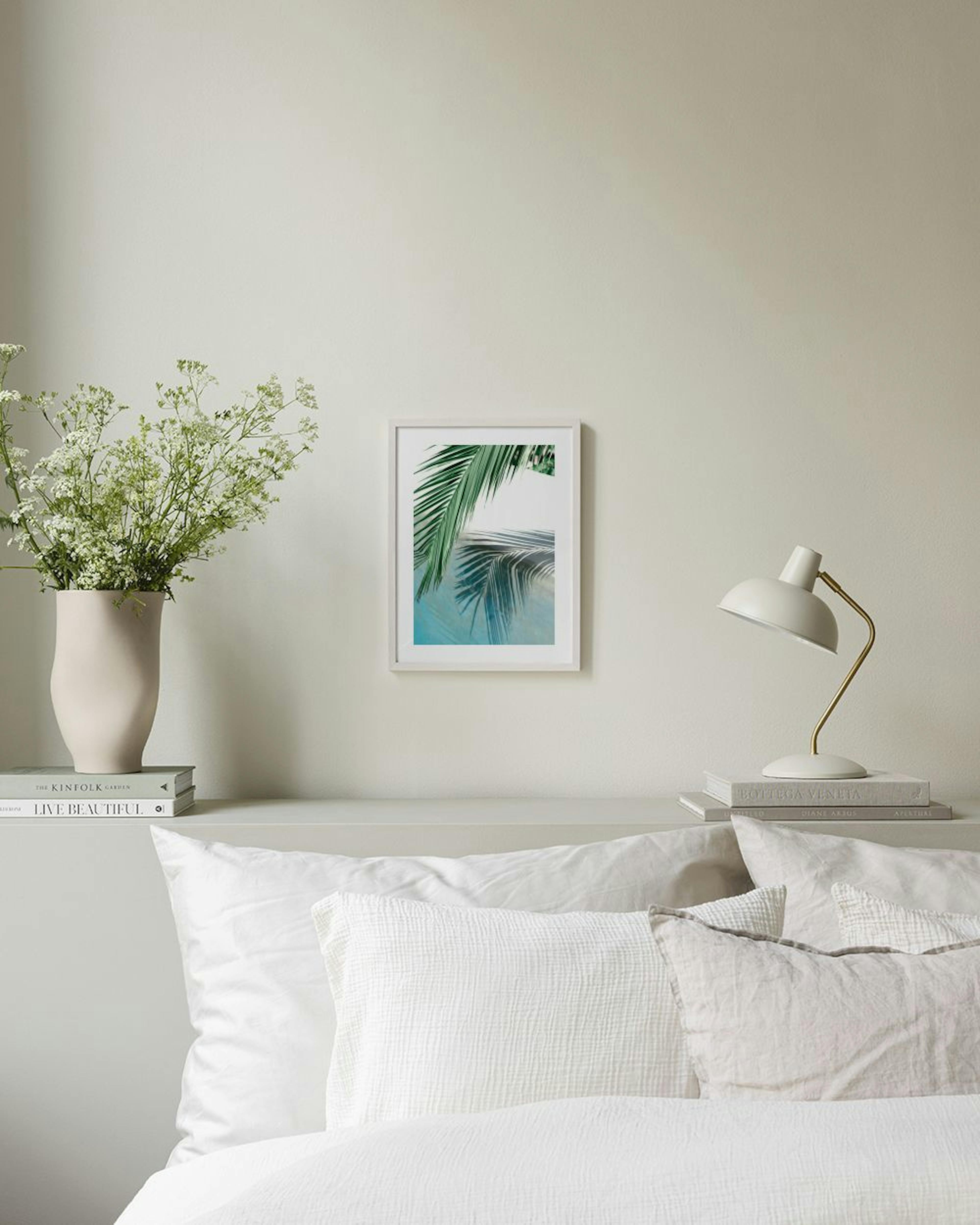 A print of green palm fronds and their reflection on a blue surface, hanging above a woven bench with books.