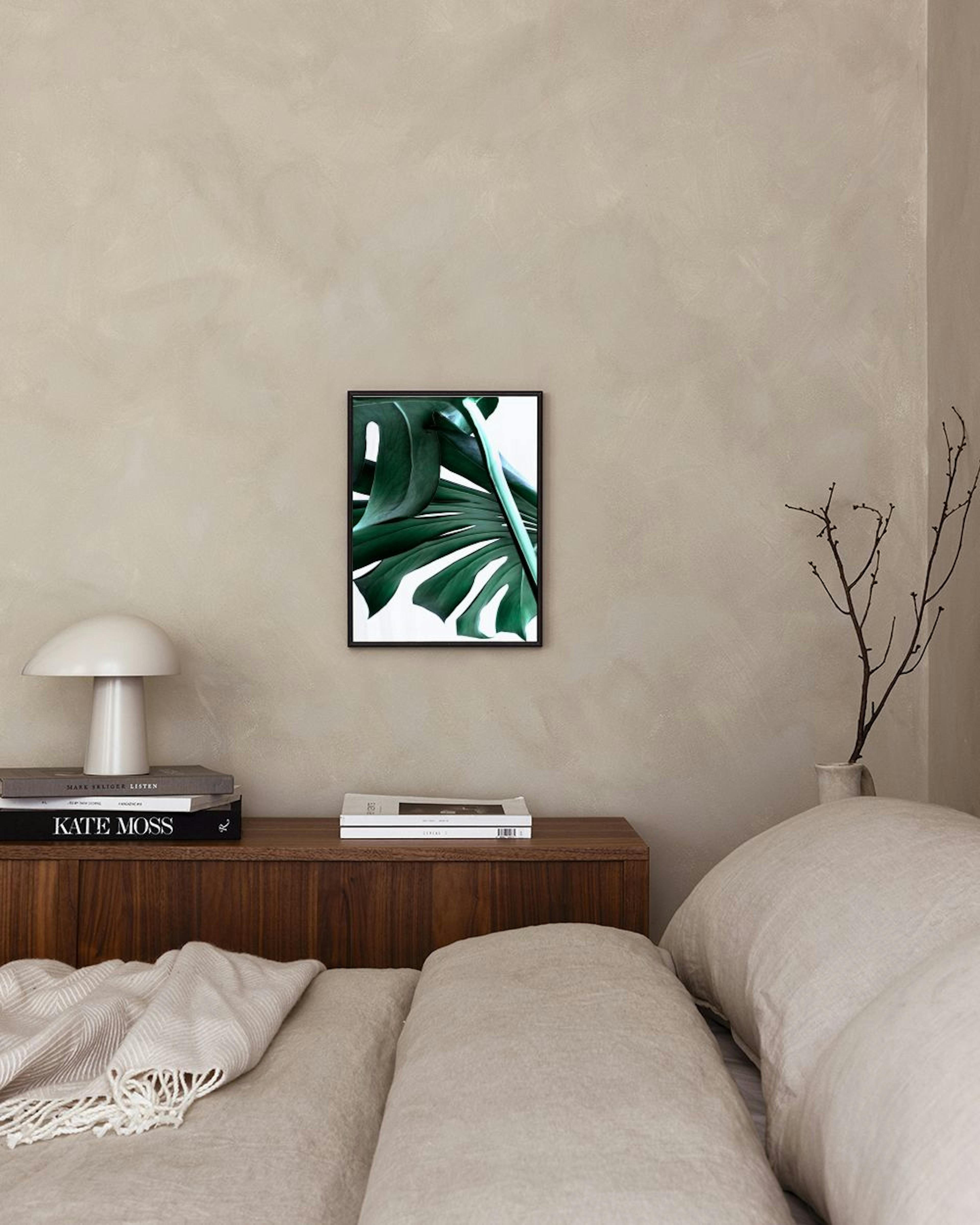 Póster de una hoja grande de monstera verde oscuro con un fondo blanco, colgado en una pared blanca sobre un taburete con libros
