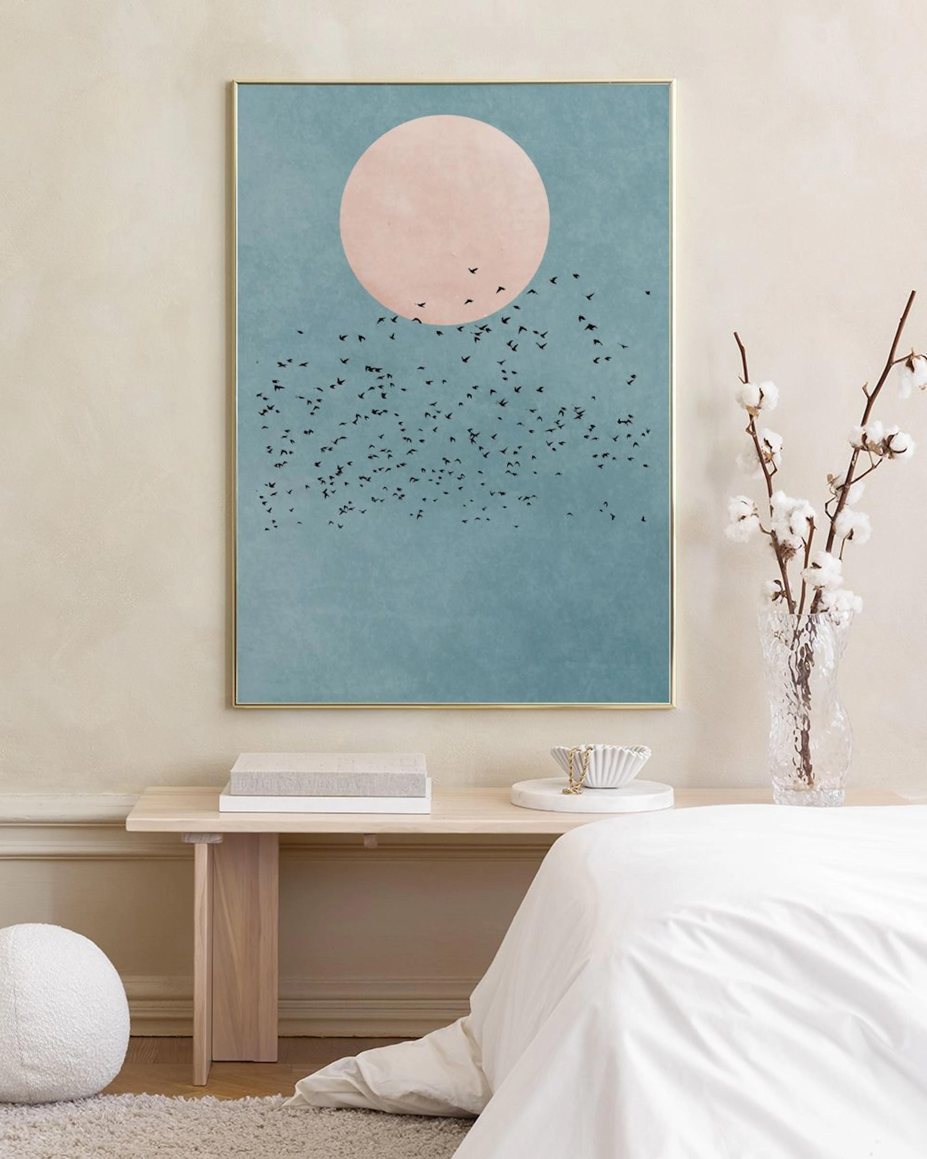 Plakat med rosa sol og flokk av fugler mot en blå himmel, henger på en hvit vegg over en krakk med bøker.