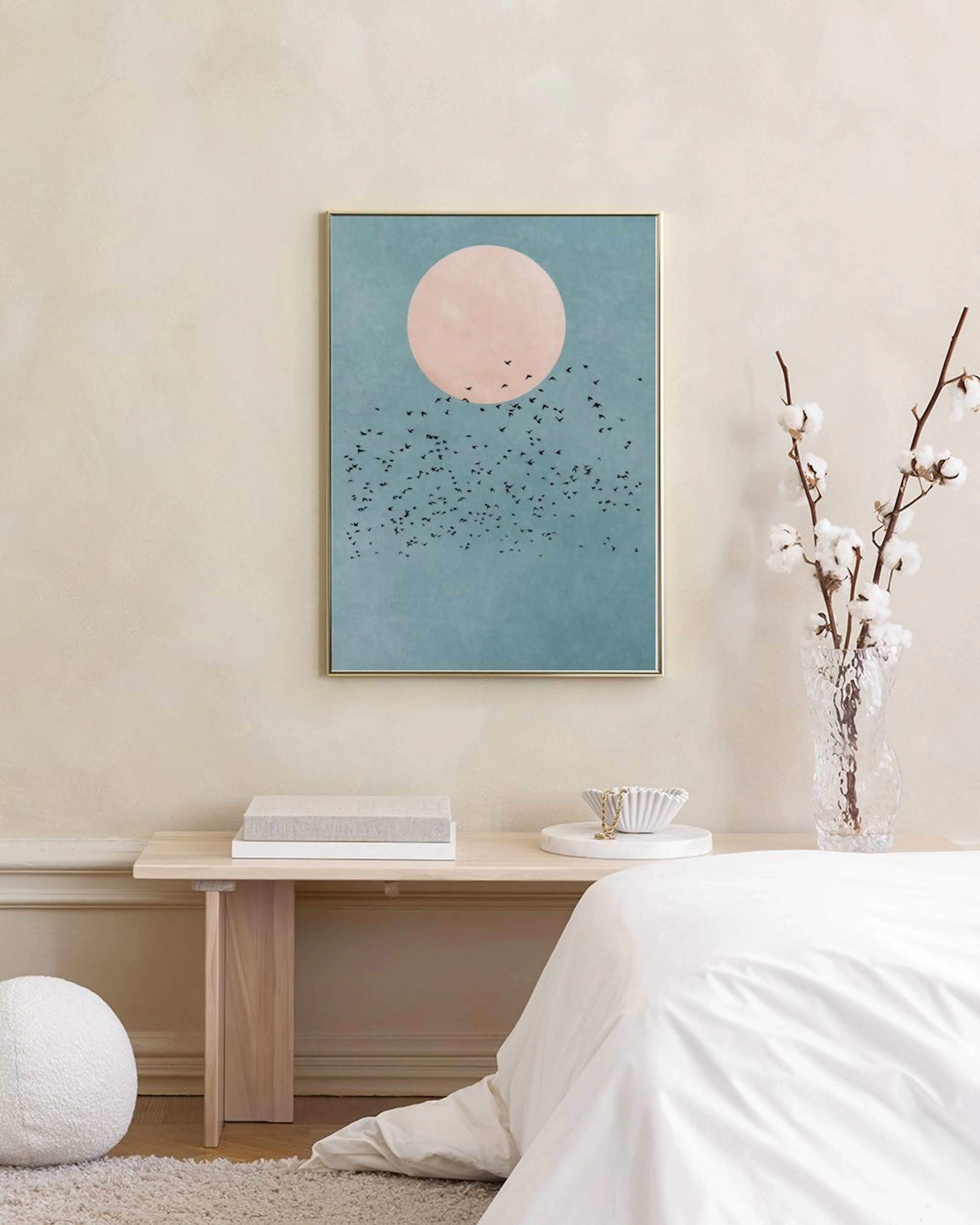 Poster med rosa sol och fågelflock mot blå himmel, hängande på vit vägg ovanför pall med böcker.
