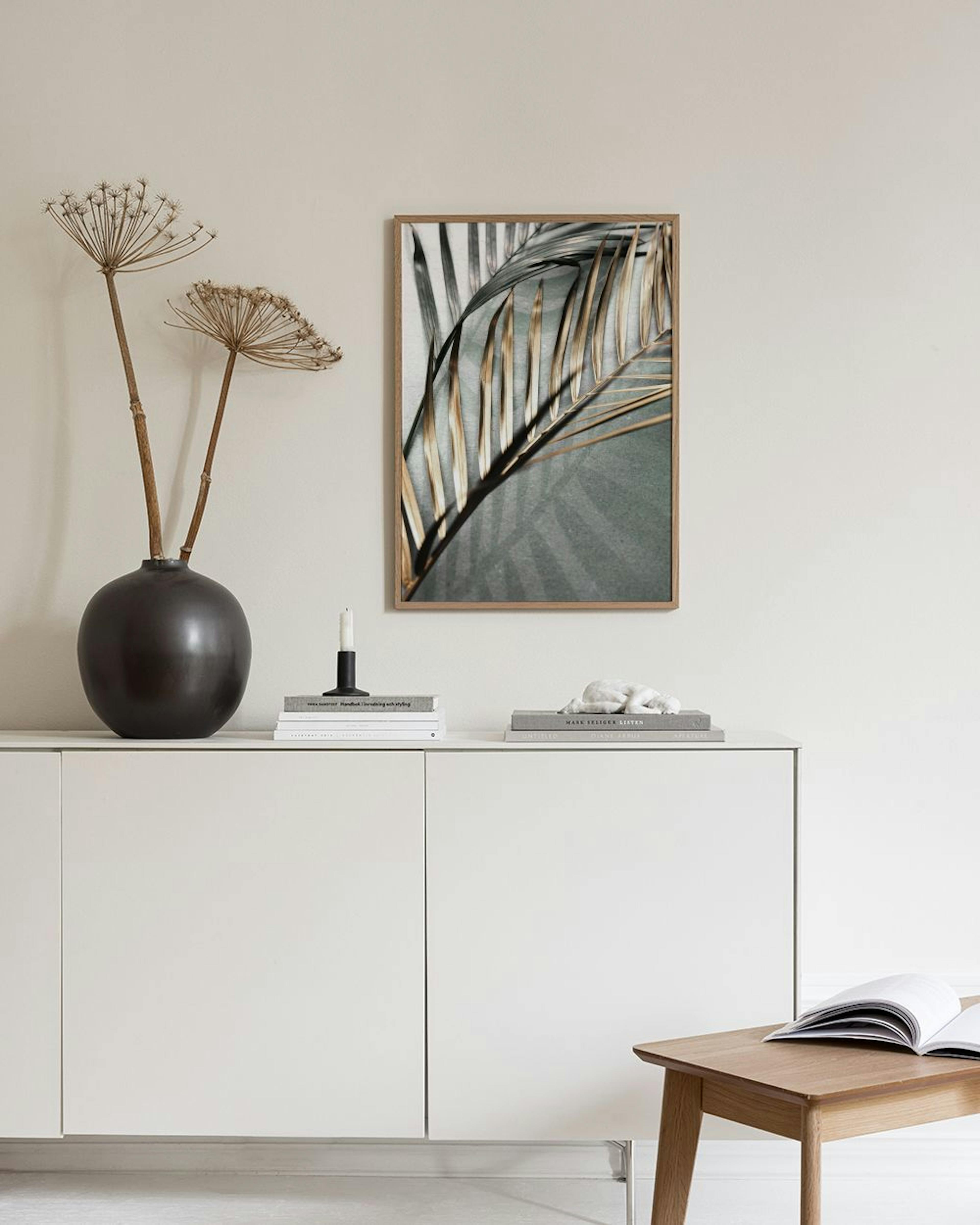 Botanical print of a dried palm leaf in gold and grey, hanging above a teal armchair in a living room.
